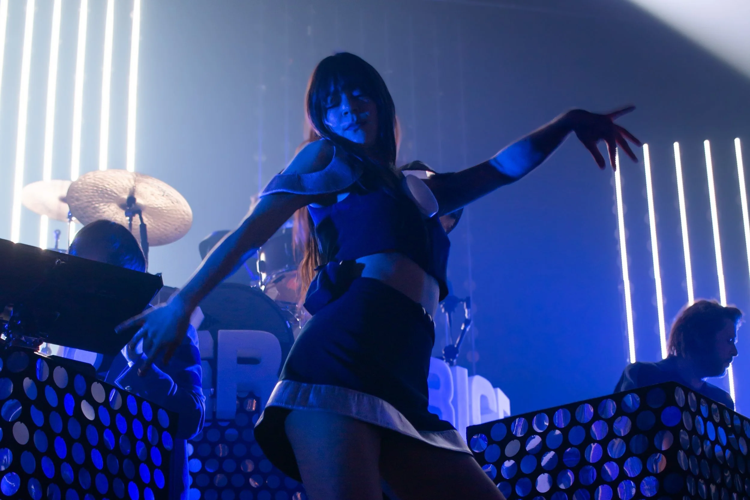 Une femme danse sur scène avec des lumières violettes et un fond de néons, accompagnée de musiciens derrière elle.