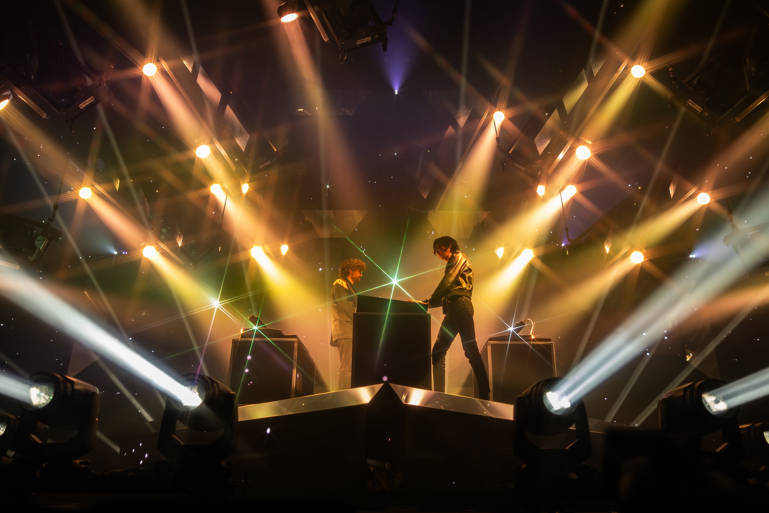 Deux DJ sur scène, illuminés par des lumières dorées et multicolores lors d'un concert, avec des effets laser et des projecteurs.