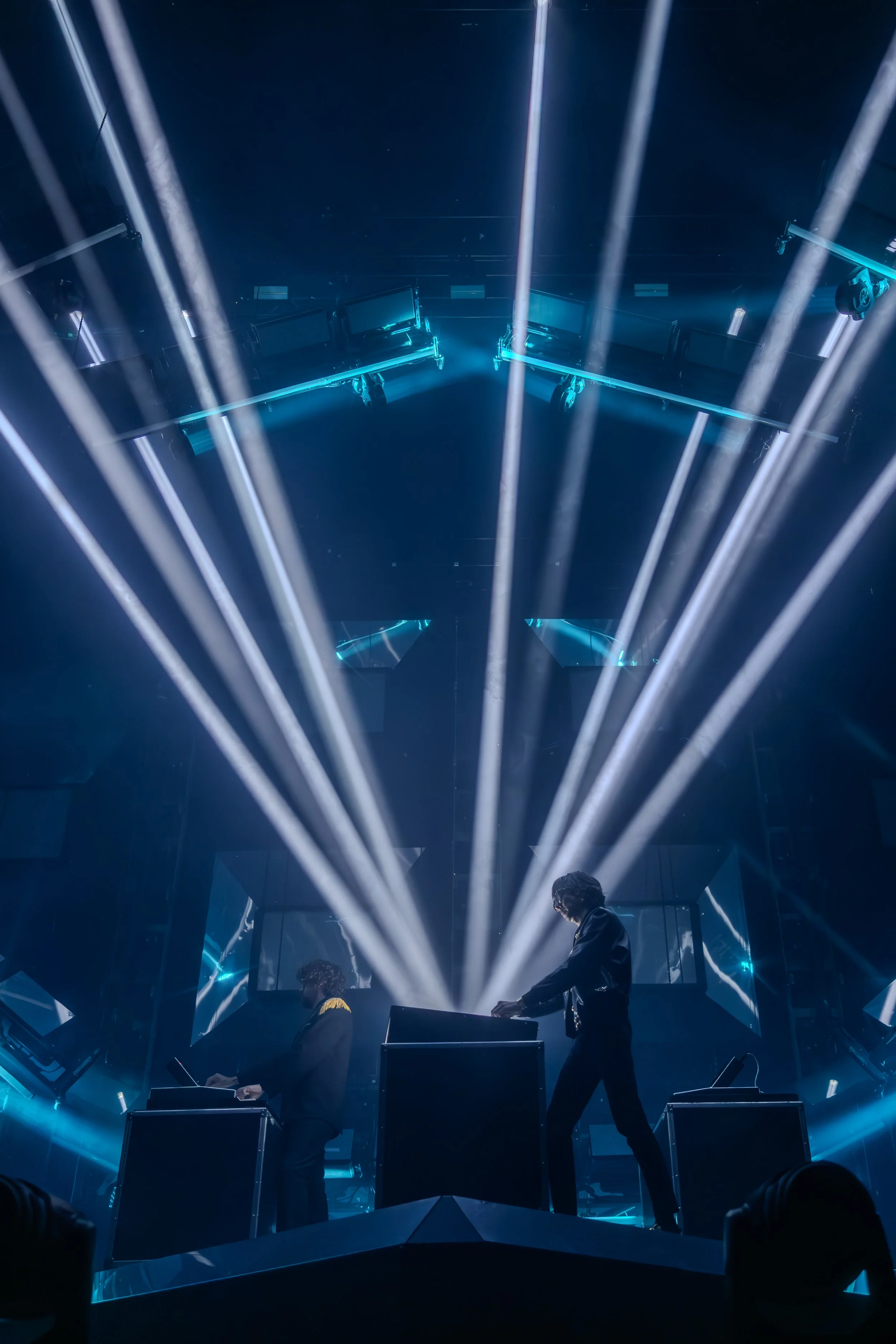 Deux DJs jouent sur scène avec des lumières laser bleues et blanches dans une ambiance de concert futuriste.