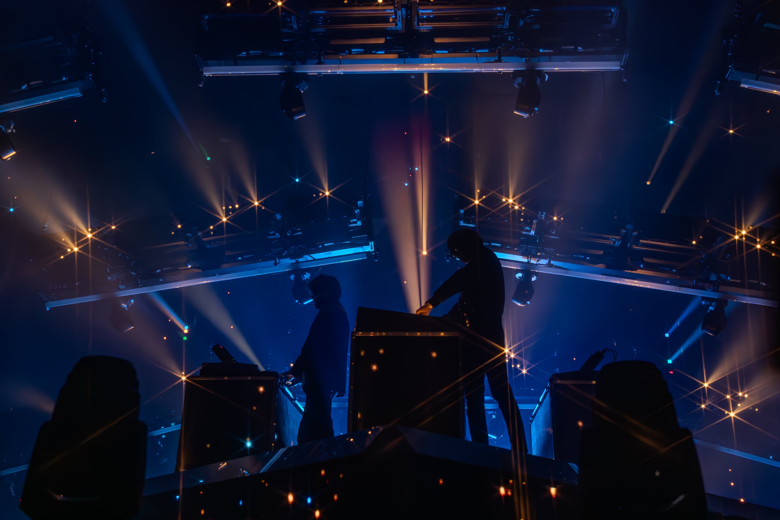 Deux DJ jouent sur une scène sombre avec des lumières bleu et orange, créant un effet lumineux étoilé.