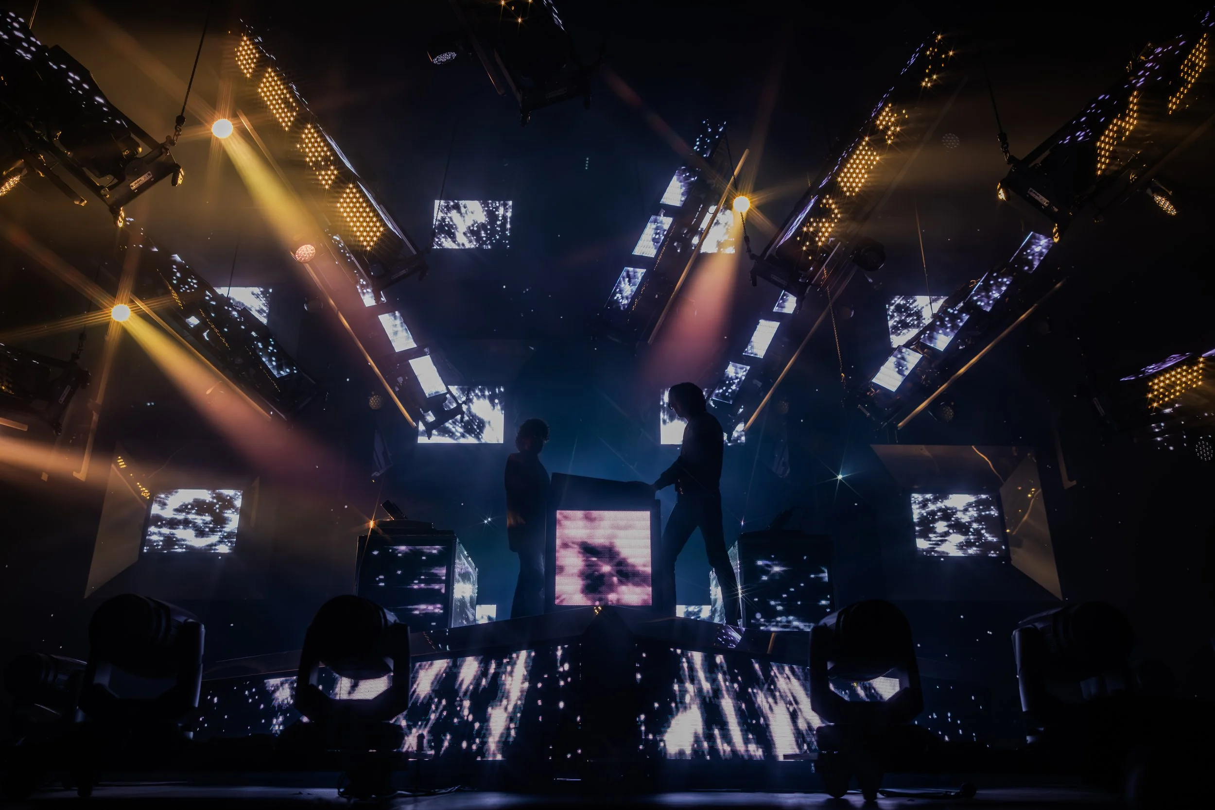 Deux personnes en silhouette jouent sur une scène de concert ou de DJ avec des lumières colorées et des écrans lumineux en arrière-plan.