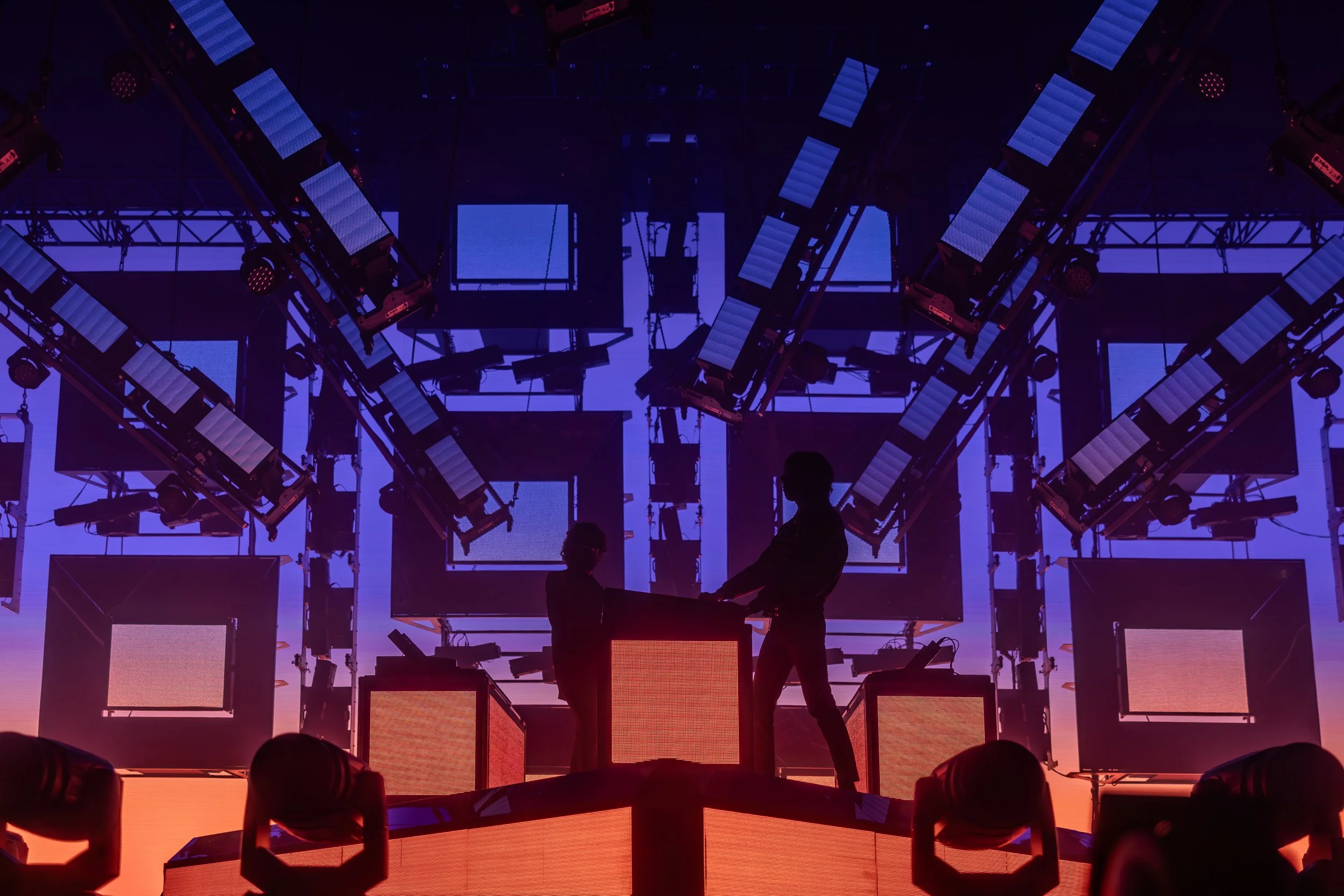 Scène de spectacle ou de concert avec silhouettes de deux personnes sur scène, entourées de lumières et d'écrans lumineux en arrière-plan, avec une ambiance colorée en violet, orange et bleu.