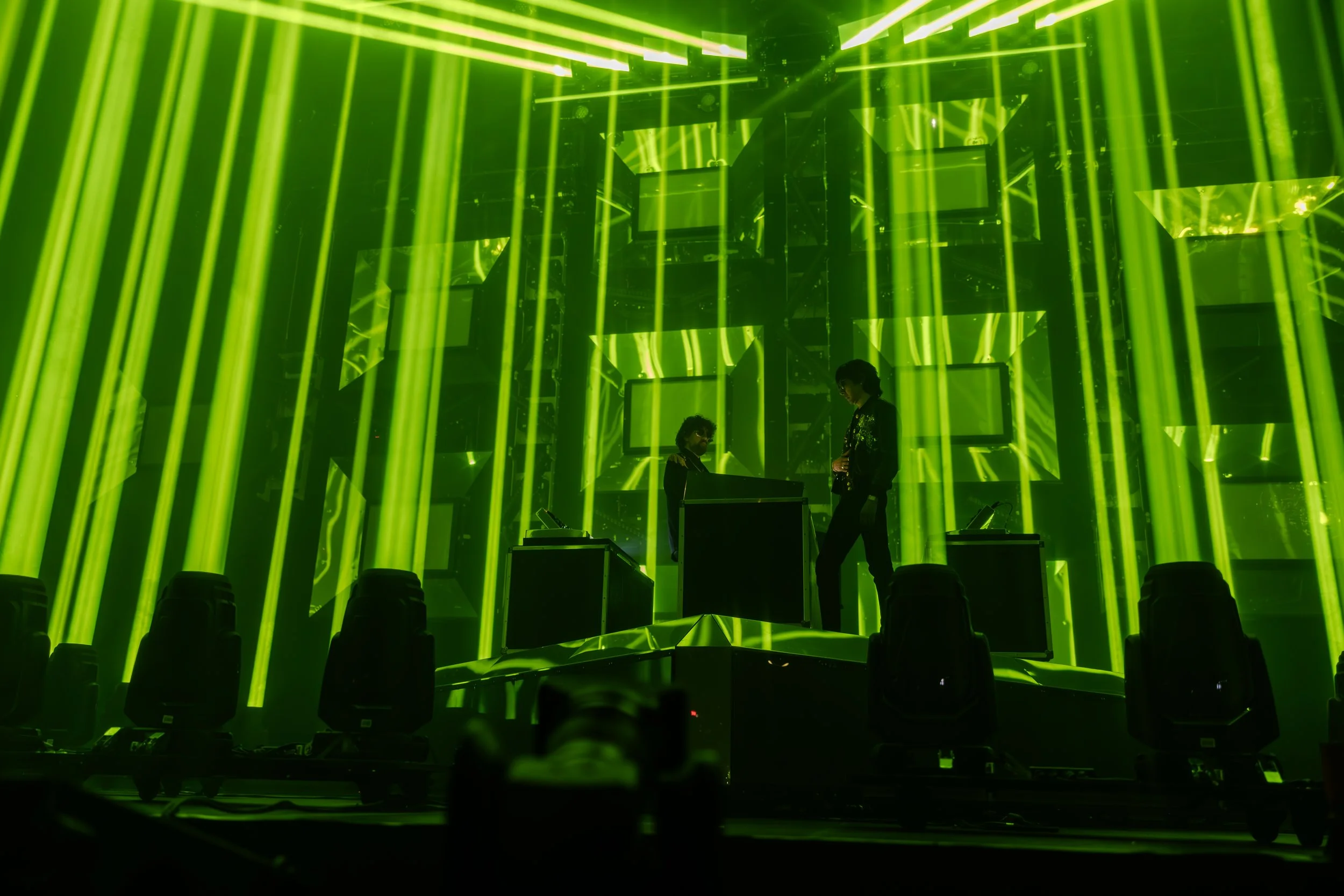 Deux personnes derrière un plateau de DJ dans une scène éclairée de rayons verts et de cadres sur un fond abstrait.