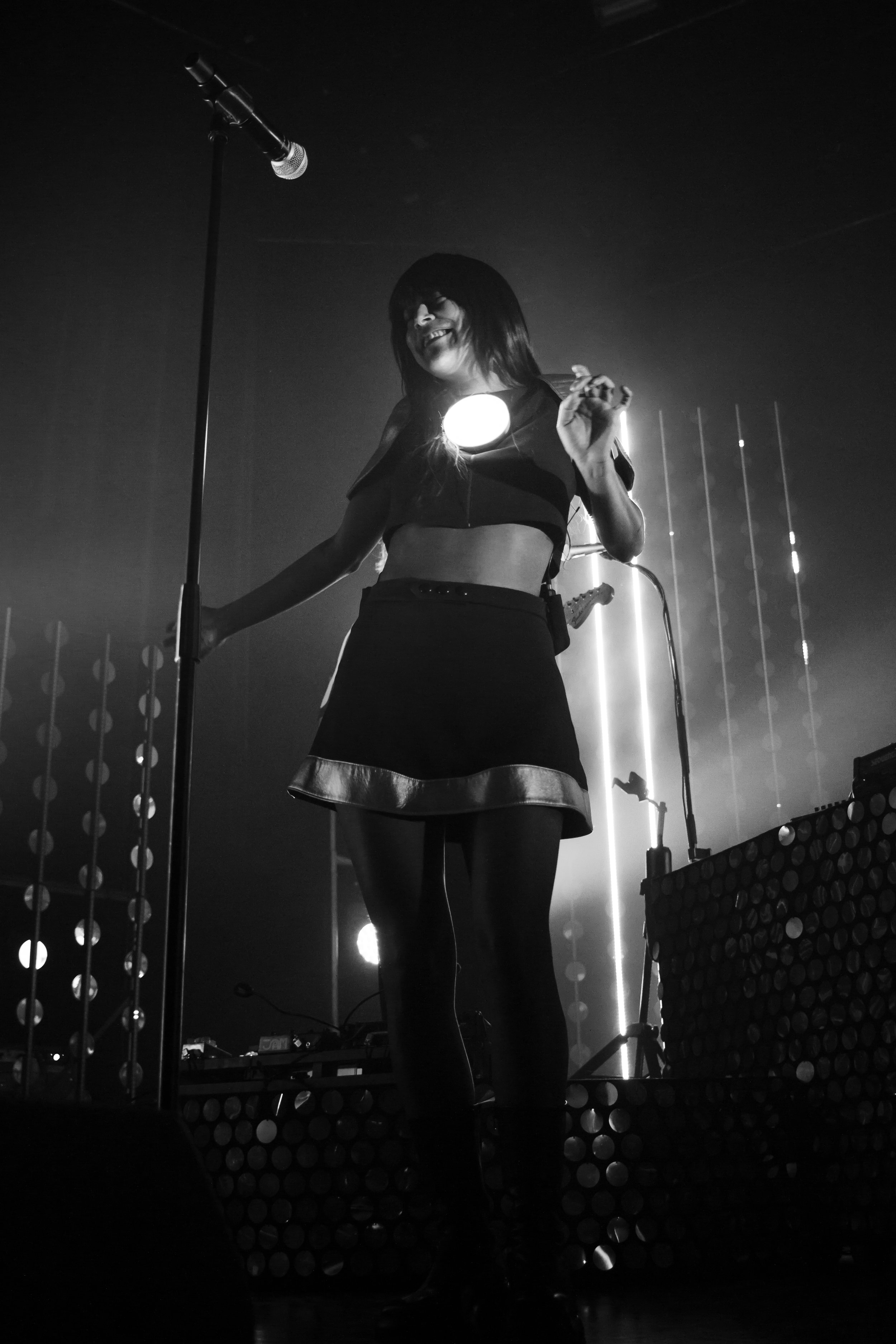 Une femme chante sur scène avec un micro, vêtue d'un haut court et d'une jupe, en noir et blanc, avec des lumières de scène derrière.