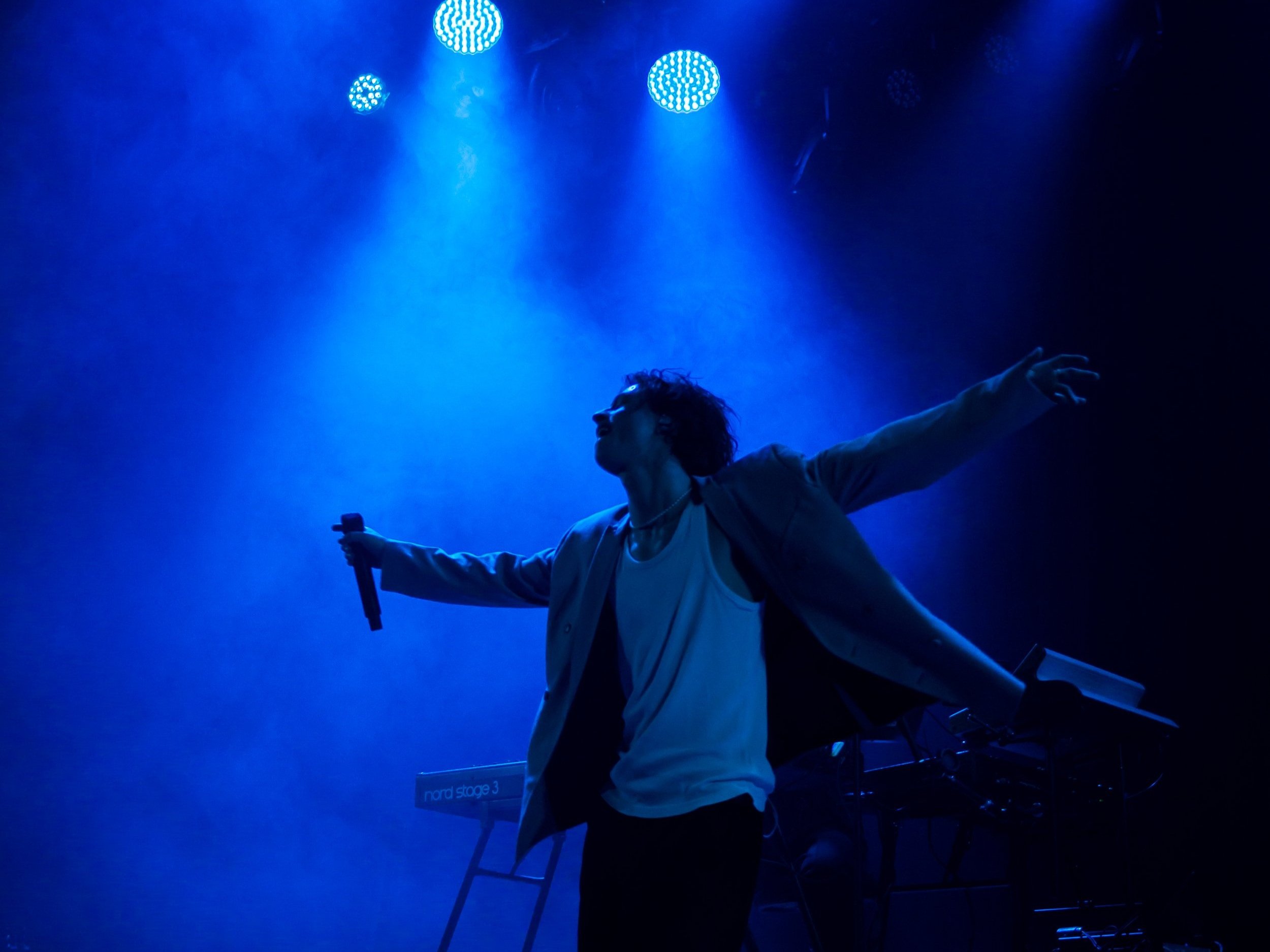 Un personnage sur scène, éclairé par des lumières bleues, avec les bras ouverts, tenant un micro dans une main. Le fond est obscur avec un clavier et des lumières d'ambiance.