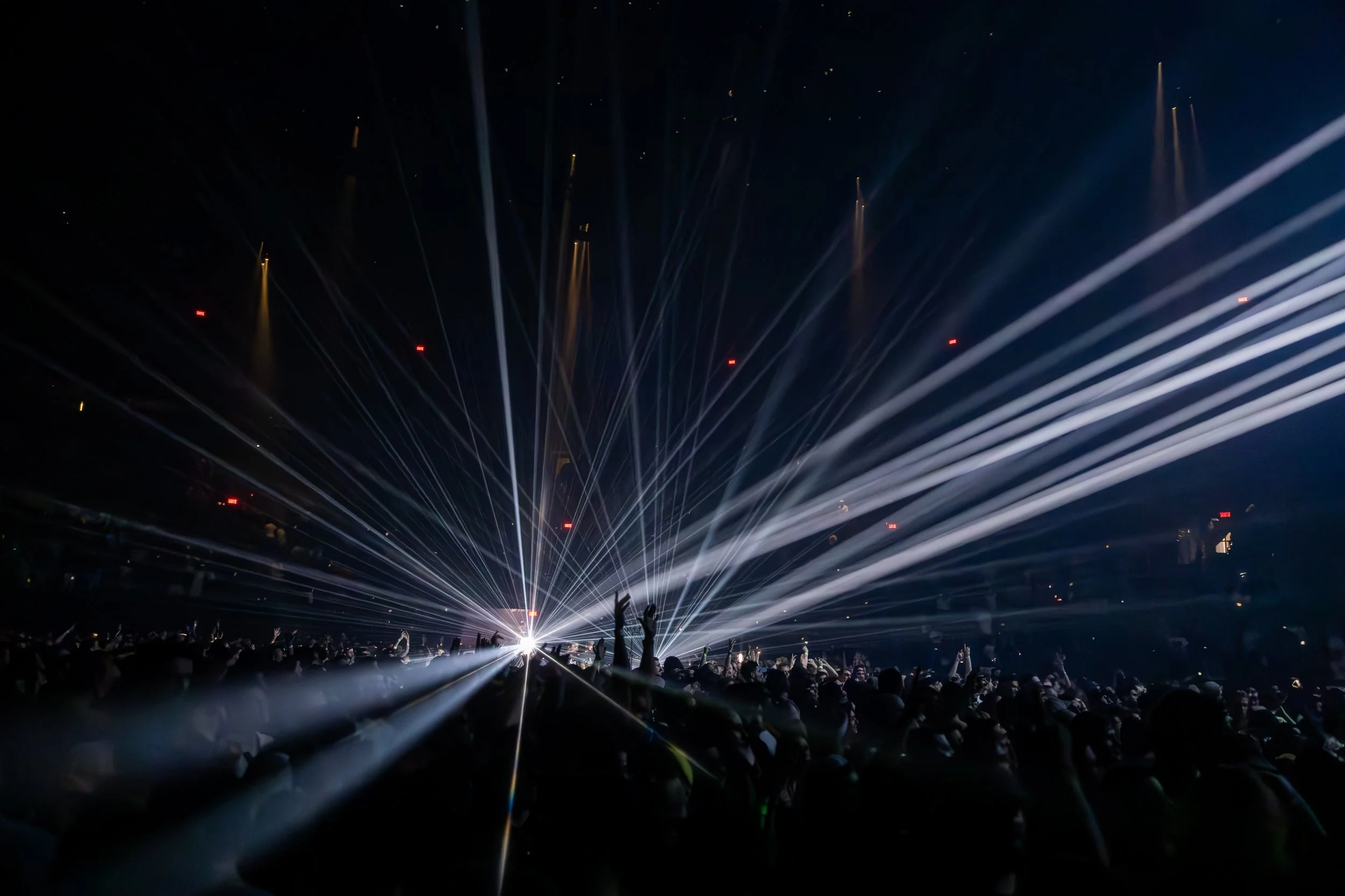 Une scène de concert avec des faisceaux lumineux blancs jaillissant du scène et un public nombreux derrière, certains avec les mains levées.