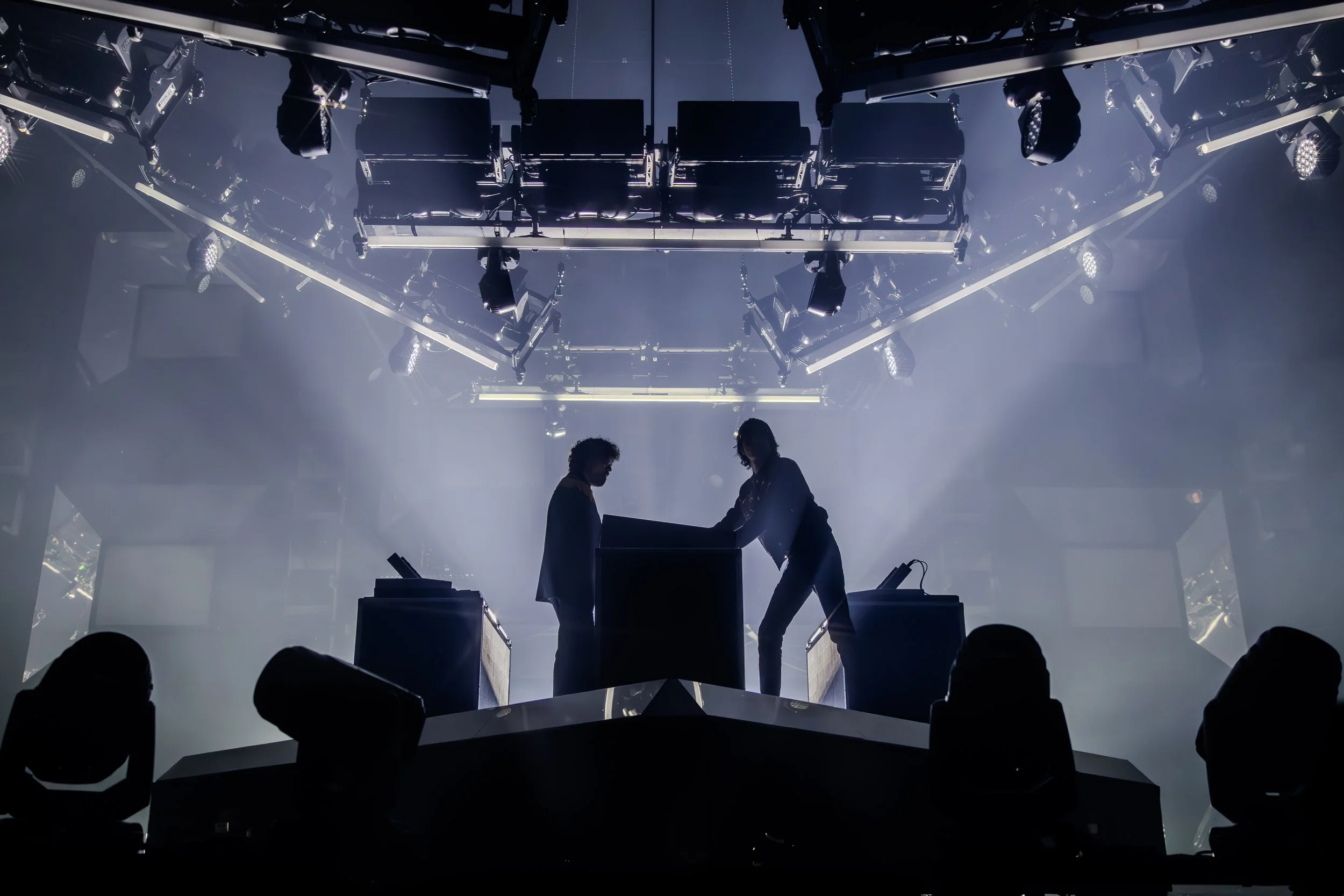 Deux personnes en silhouette sur scène avec des équipements de festival ou de concert, ambiance sombre et éclairage dramatique, avec des lumières suspendues au plafond.