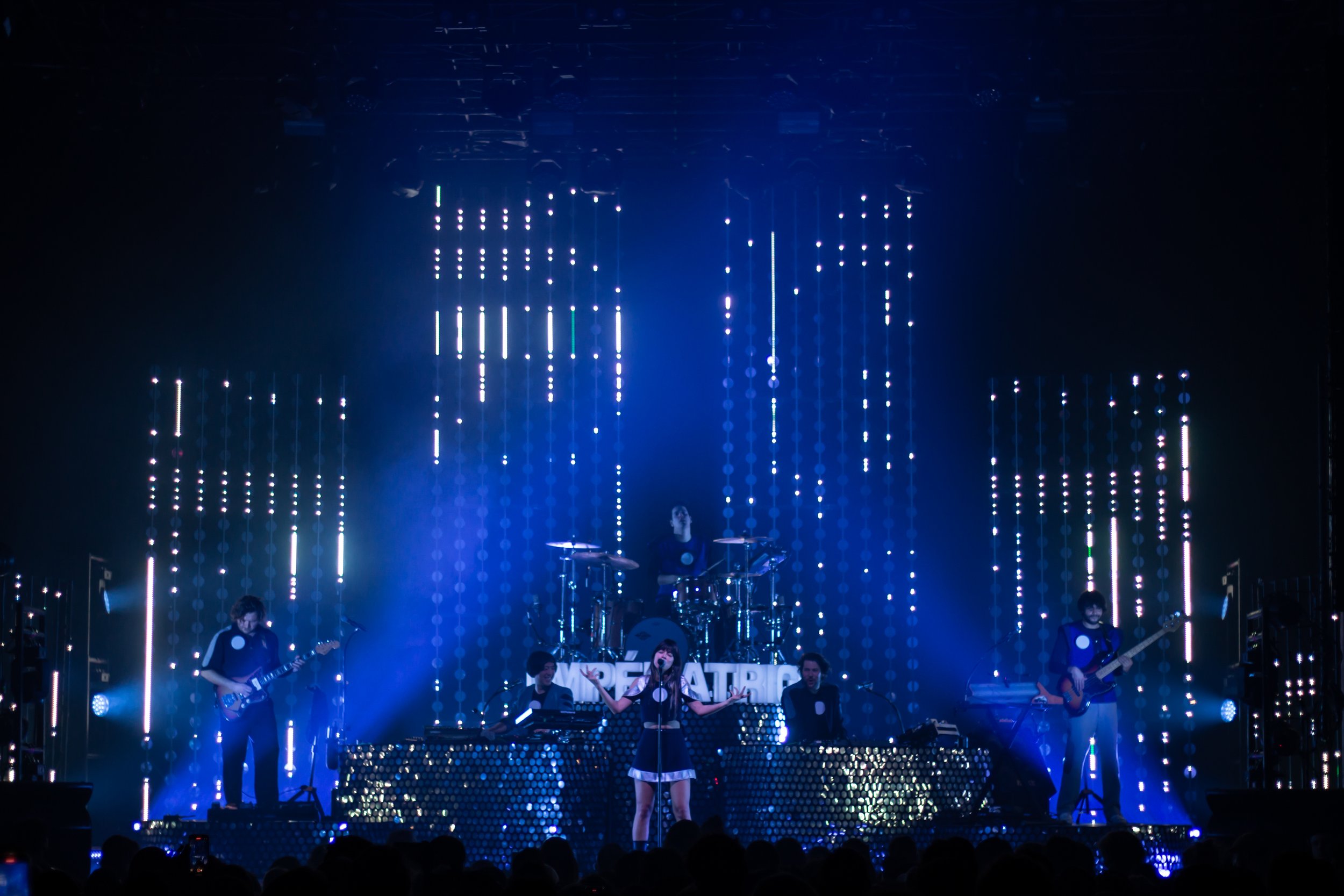 Une performance musicale sur scène avec un groupe de cinq musiciens, éclairage bleu, arrière-plan avec des lumières verticales LED et écran affichant le mot 'DEATRA.'