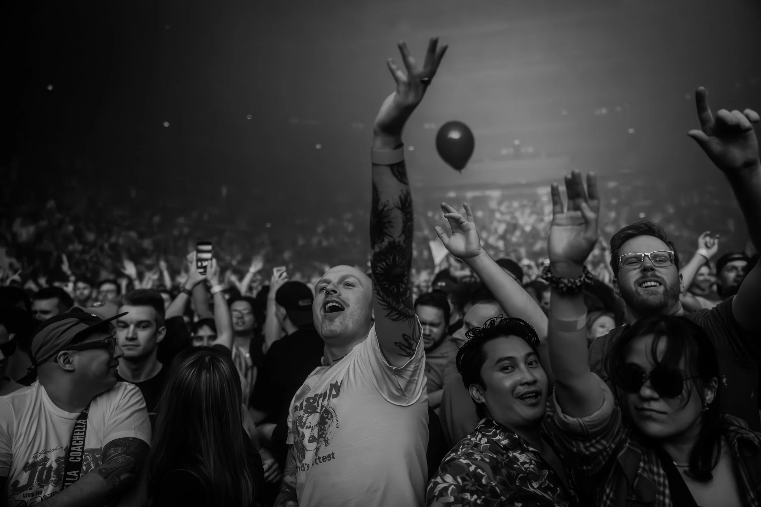 Une foule de personnes souriantes et joyeuses lors d'un concert ou festival, avec des bras levés, dans une ambiance énergique, en noir et blanc.