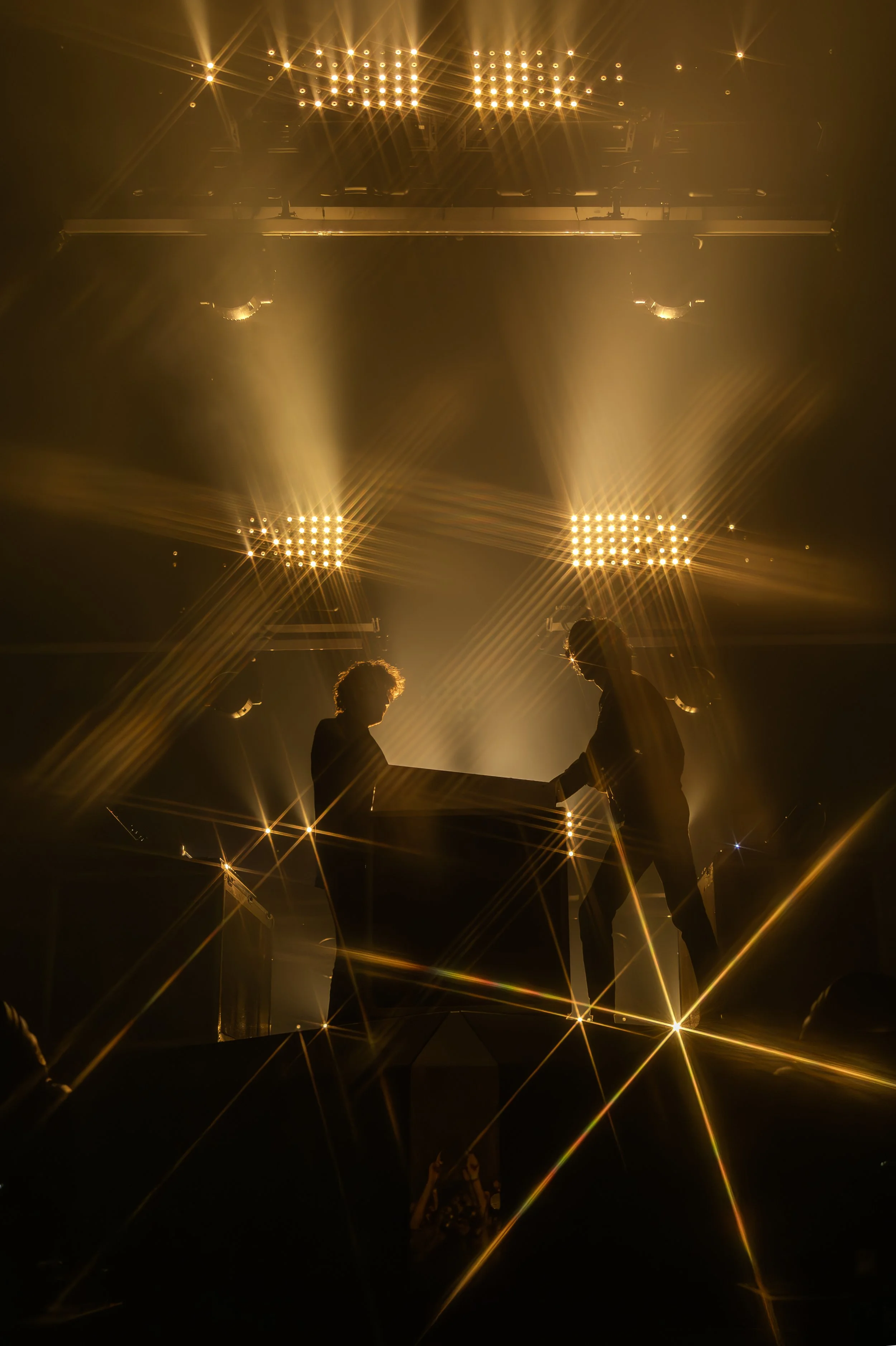 Deux personnes en silhouette échangeant un objet ou une main lors d'une scène mise en valeur par un éclairage doré et brillant, avec des lumières armés au plafond créant un effet de brillance et de reflets dans l'obscurité.