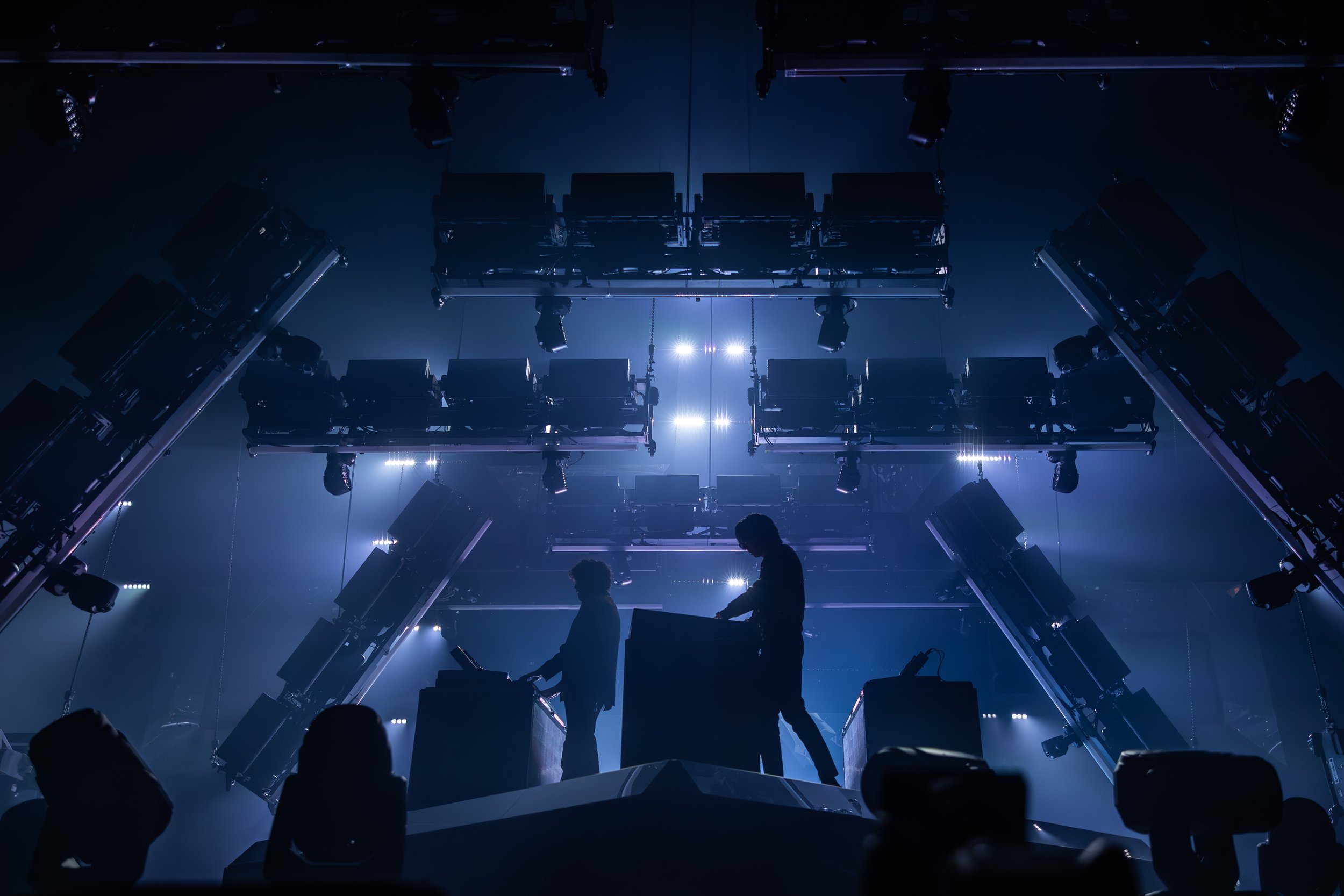Deux DJ en silhouette sur une scène illuminée de lumières bleues, entourés de nombreux équipements de son et de lumières suspendues au plafond.