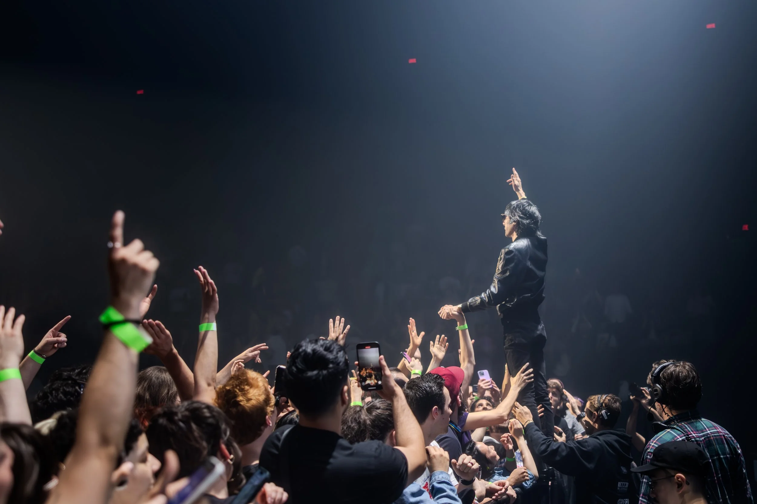 Une personne se tient au-dessus d'une foule lors d'un concert ou d'un événement, le bras levé, sous une lumière tamisée.