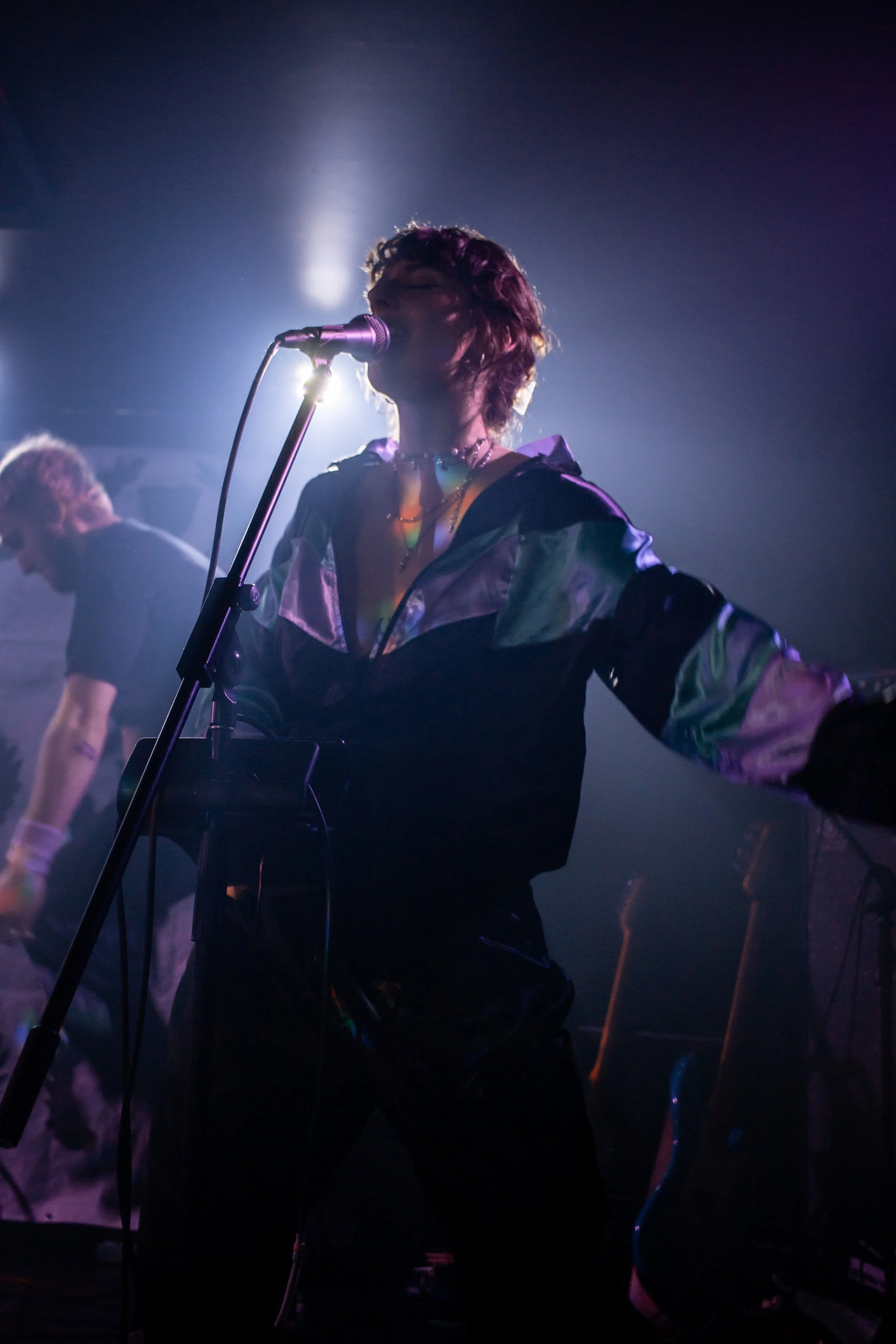Une femme chante en microphone lors d'une performance en concert, avec un éclairage dramatique et une ambiance sombre, accompagnée d'un musicien en arrière-plan.