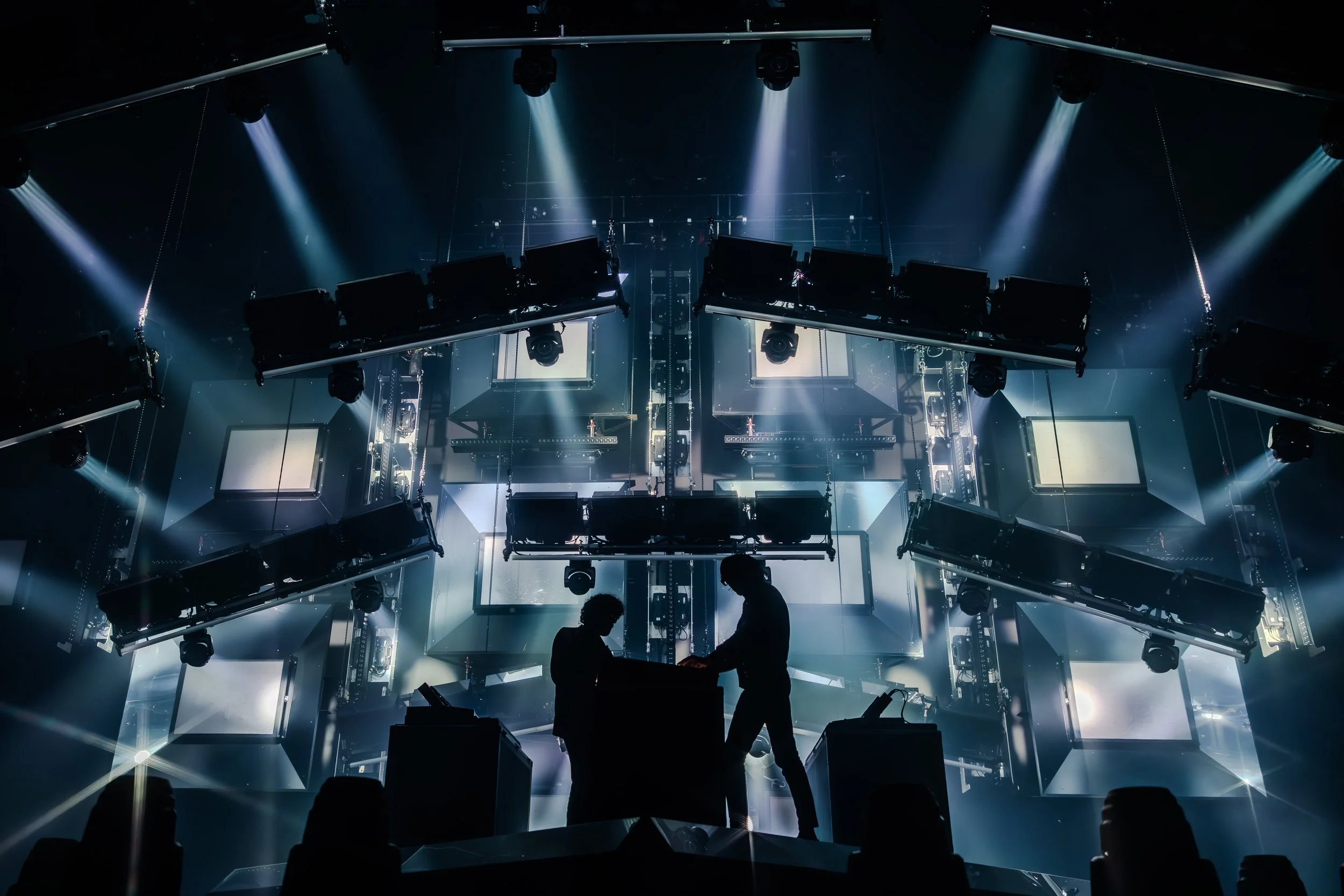 Silhouettes de deux personnes travaillant sur scène avec un décor de lumières et d'écrans lumineux en arrière-plan, créant un effet dramatique et futuriste.