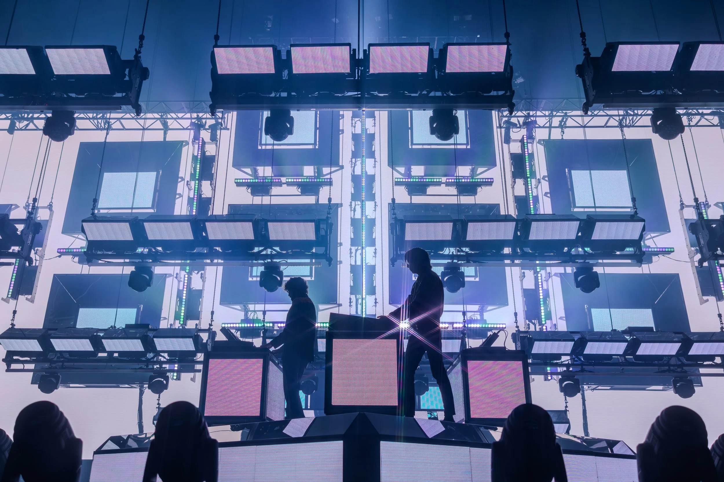 Deux personnes en silhouette jouent sur une scène de concert lumineuse avec des écrans LED et des lumières colorées.