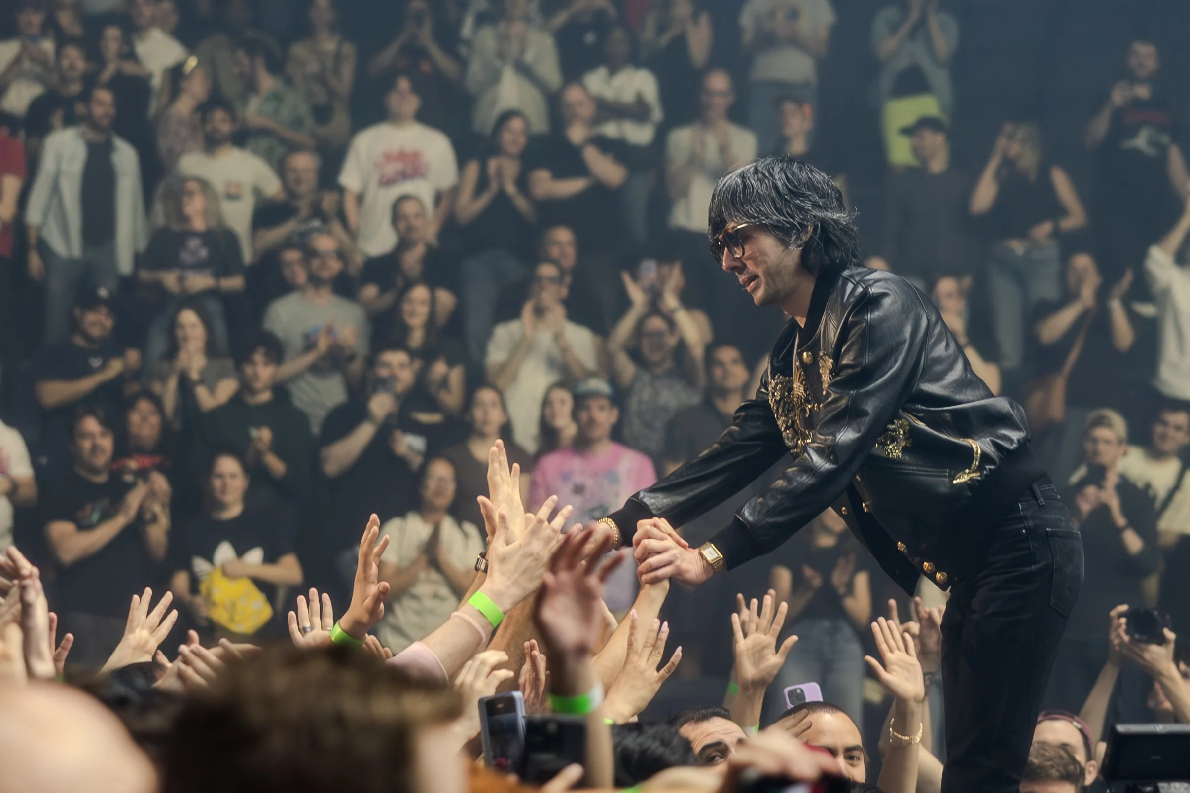 Un homme en vêtements noirs et lunettes, avec des cheveux gris, tend la main pour aider un membre du public lors d'un concert ou spectacle, avec une foule applaudissant en arrière-plan.