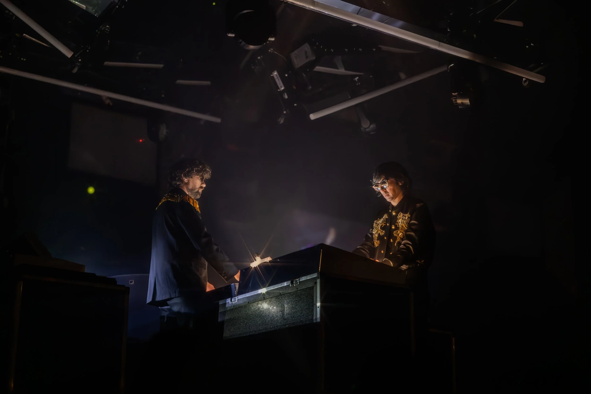 Deux musiciens jouent du synthétiseur dans une scène sombre, éclairés doucement par une lumière, un homme à gauche et une femme à droite portant des vêtements ornés de broderies dorées.