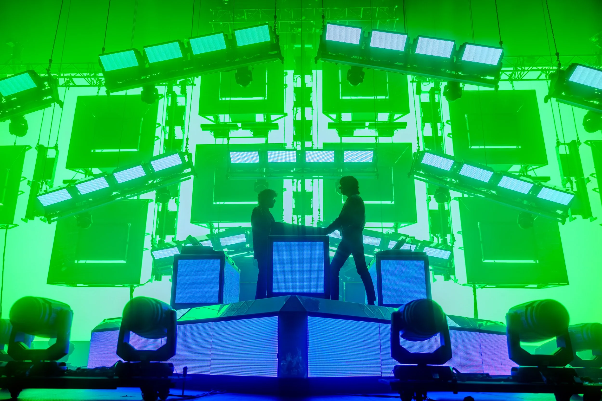 Deux personnes s'échangent un document sur une plateforme de scène avec de nombreux écrans et éclairages LED en arrière-plan, dans une configuration de production d'événement ou de concert.