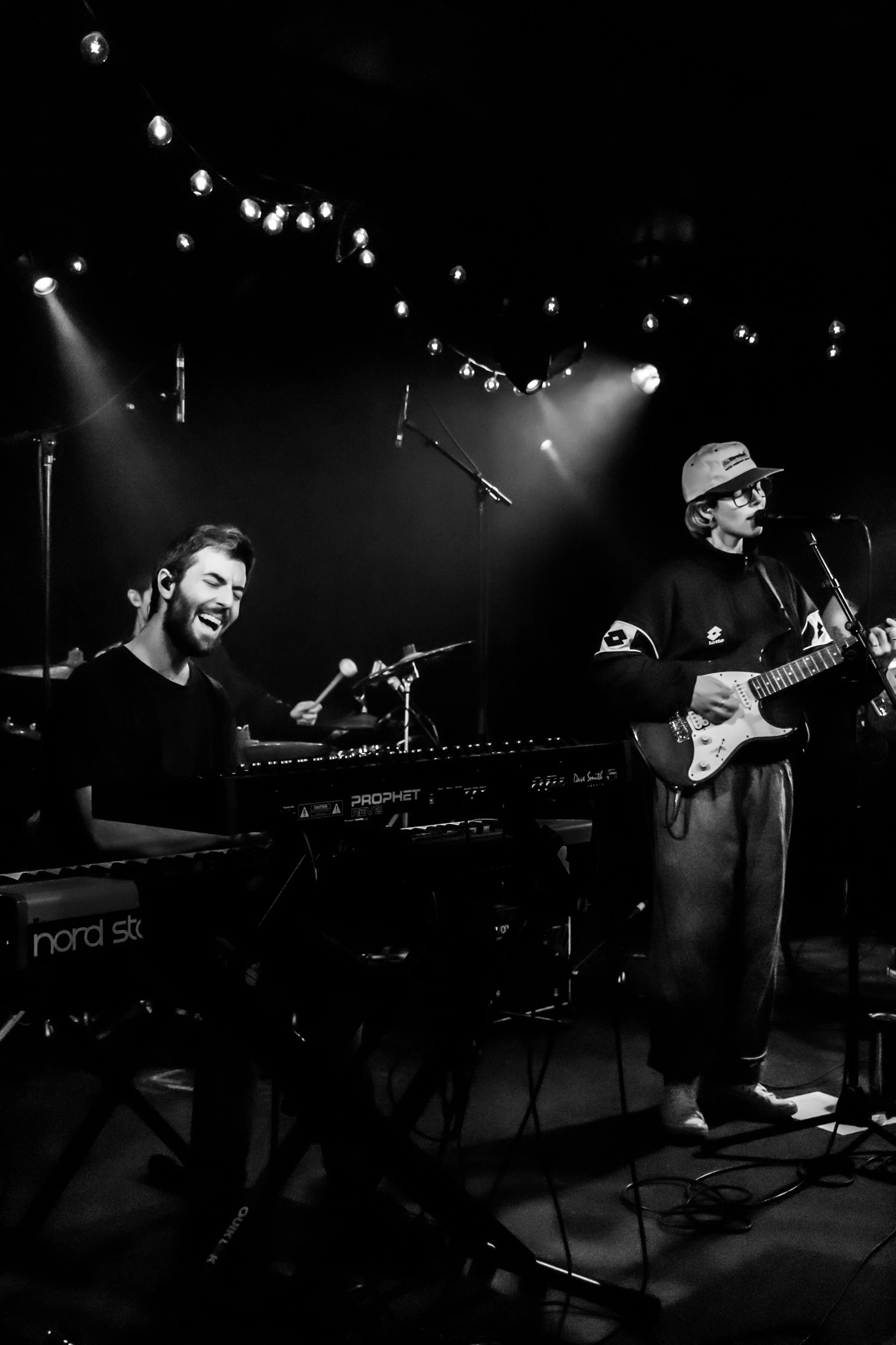 Deux musiciens en performance sur scène, un joueur de clavier souriant et un guitariste chantant, dans un environnement sombre avec des lumières et des décorations suspendues