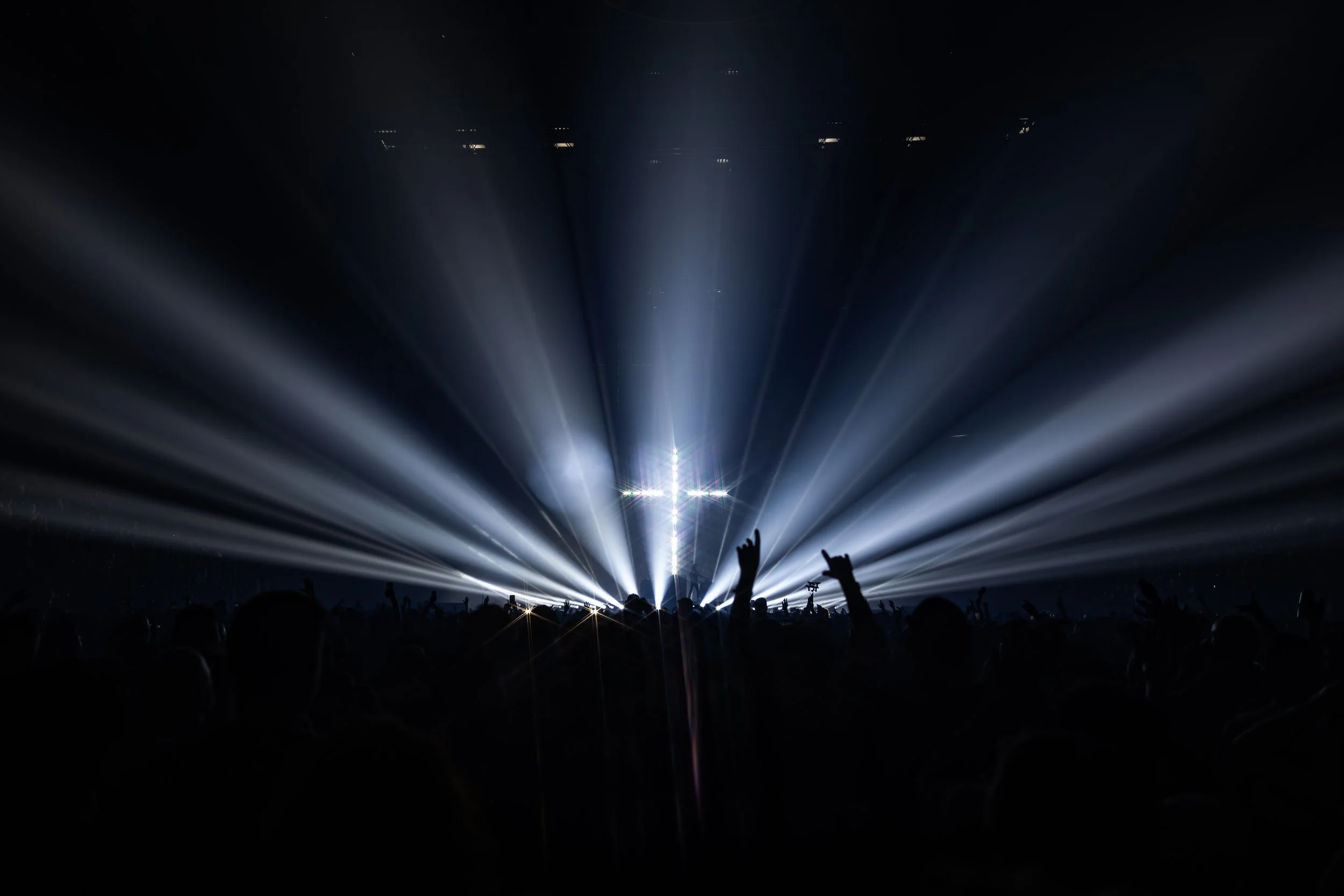 Spectacle lumineux avec des rayons lumineux bleus pointant vers le ciel lors d'un concert, silhouette de public avec des mains levées dans une ambiance sombre.