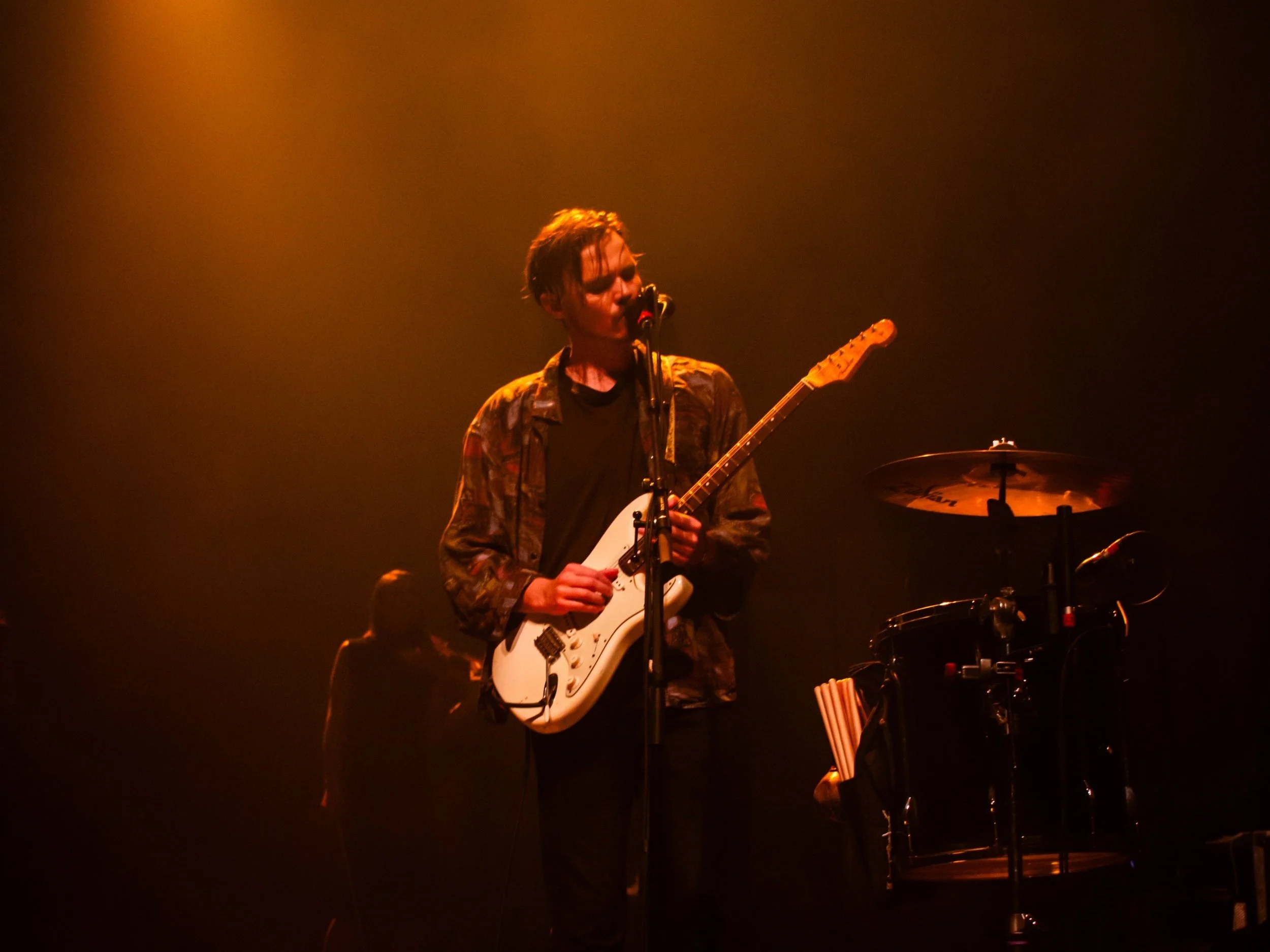 Un musicien joue de la guitare électrique lors d'un concert, éclairé par une lumière chaude, avec un autre musicien en arrière-plan.