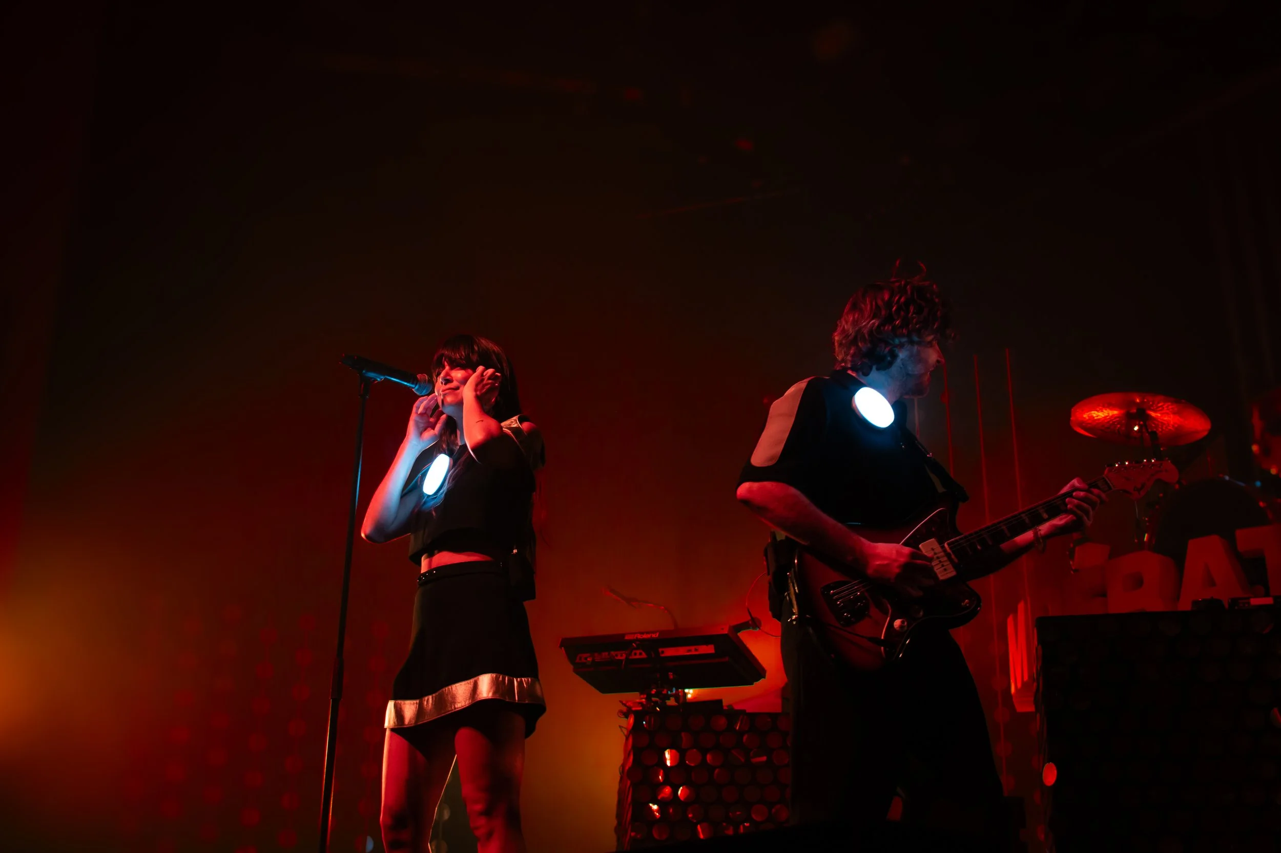 Deux musiciens sur scène, une femme chante en tenant un micro, un homme joue de la guitare électriques, scène sombre avec éclairages rouge et orange.