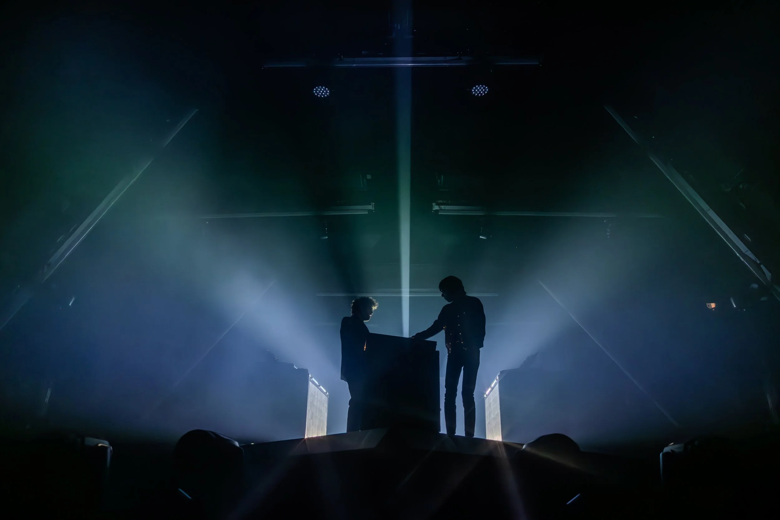 Deux personnes en silhouette jouent dans une ambiance sombre et lumineuse sur scène avec des effets de lumière bleue et blanche, semblant être des DJ ou des artistes en performance