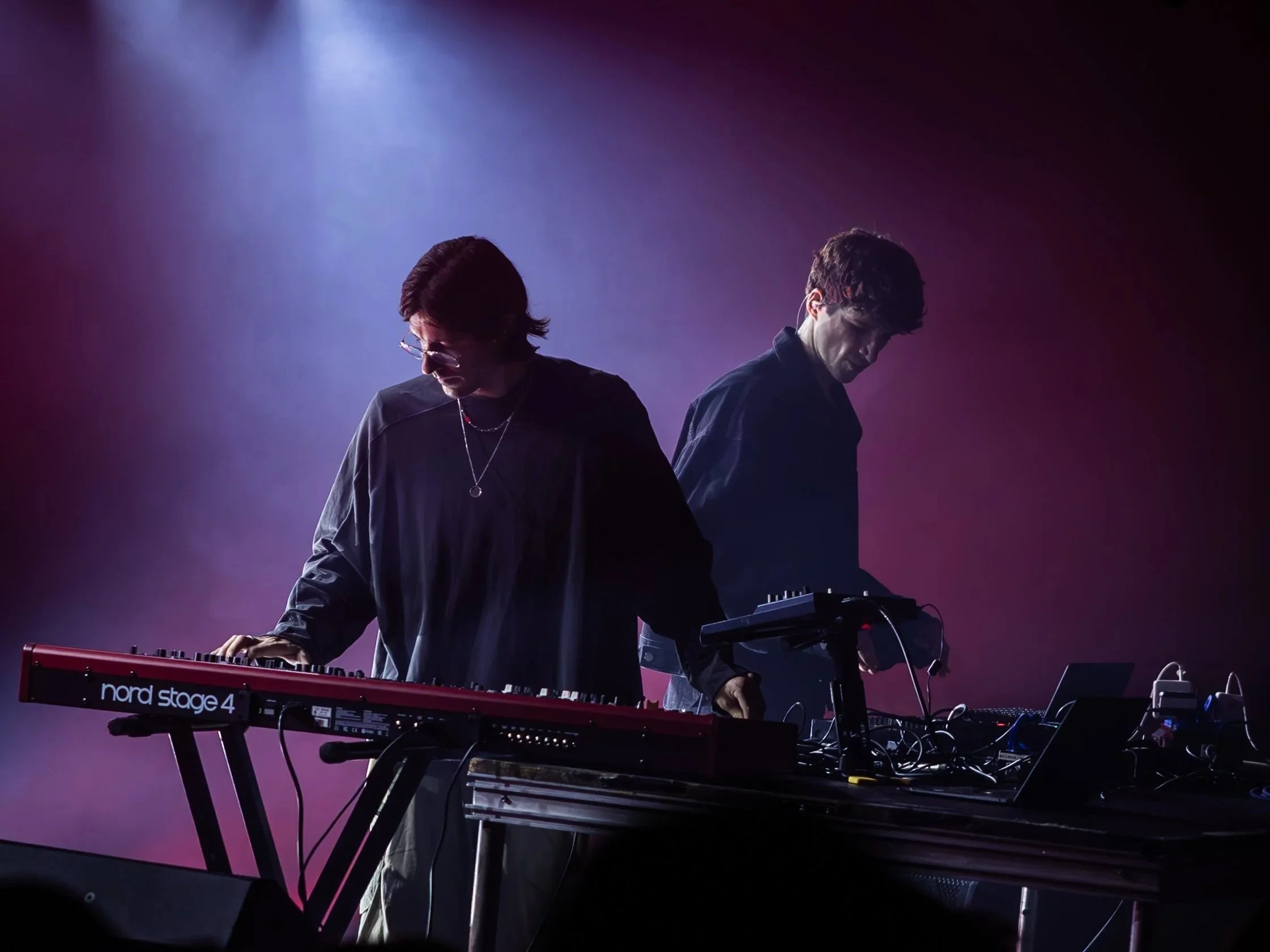 Deux musiciens jouent sur scène avec un clavier Nord Stage 4, éclairés par une lumière violette, en arrière-plan sombre.