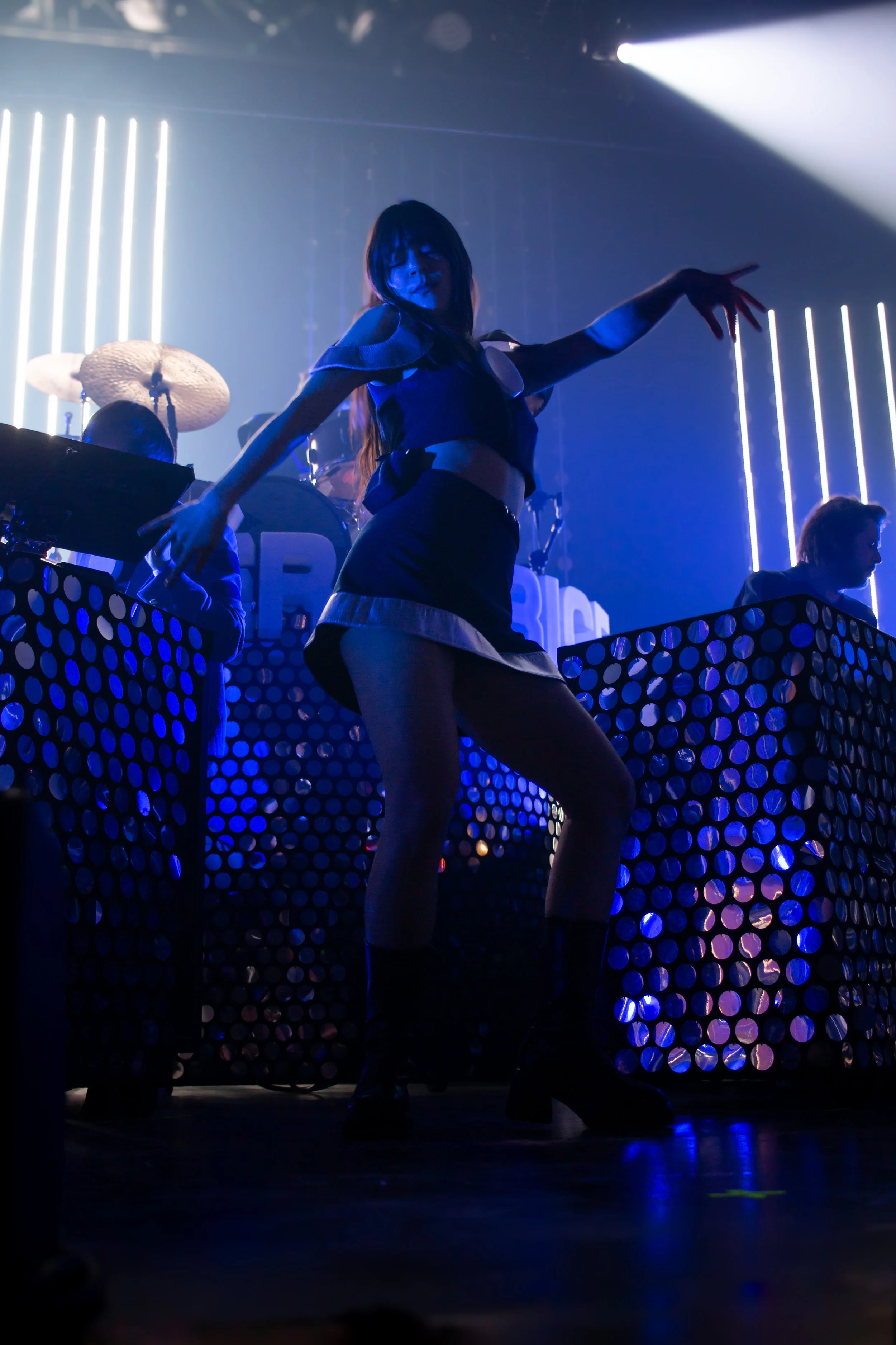 Une femme danse sur scène dans un environnement de fête ou de concert, avec des jeux de lumières bleues et des musiciens en arrière-plan.
