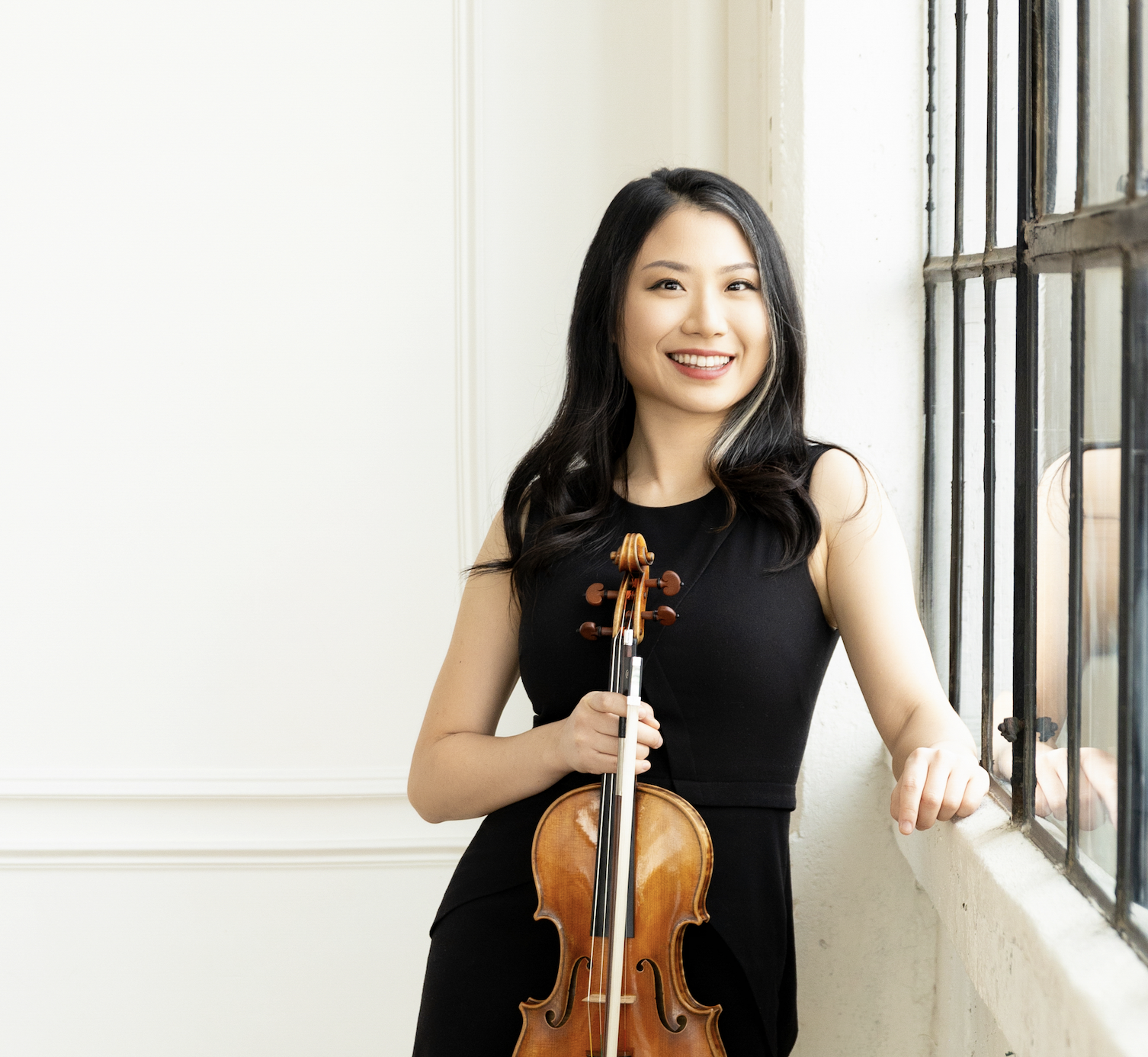 A young woman with black hair, wearing a black sleeveless dress, smiling and holding a violin, standing by a large window with metal bars.