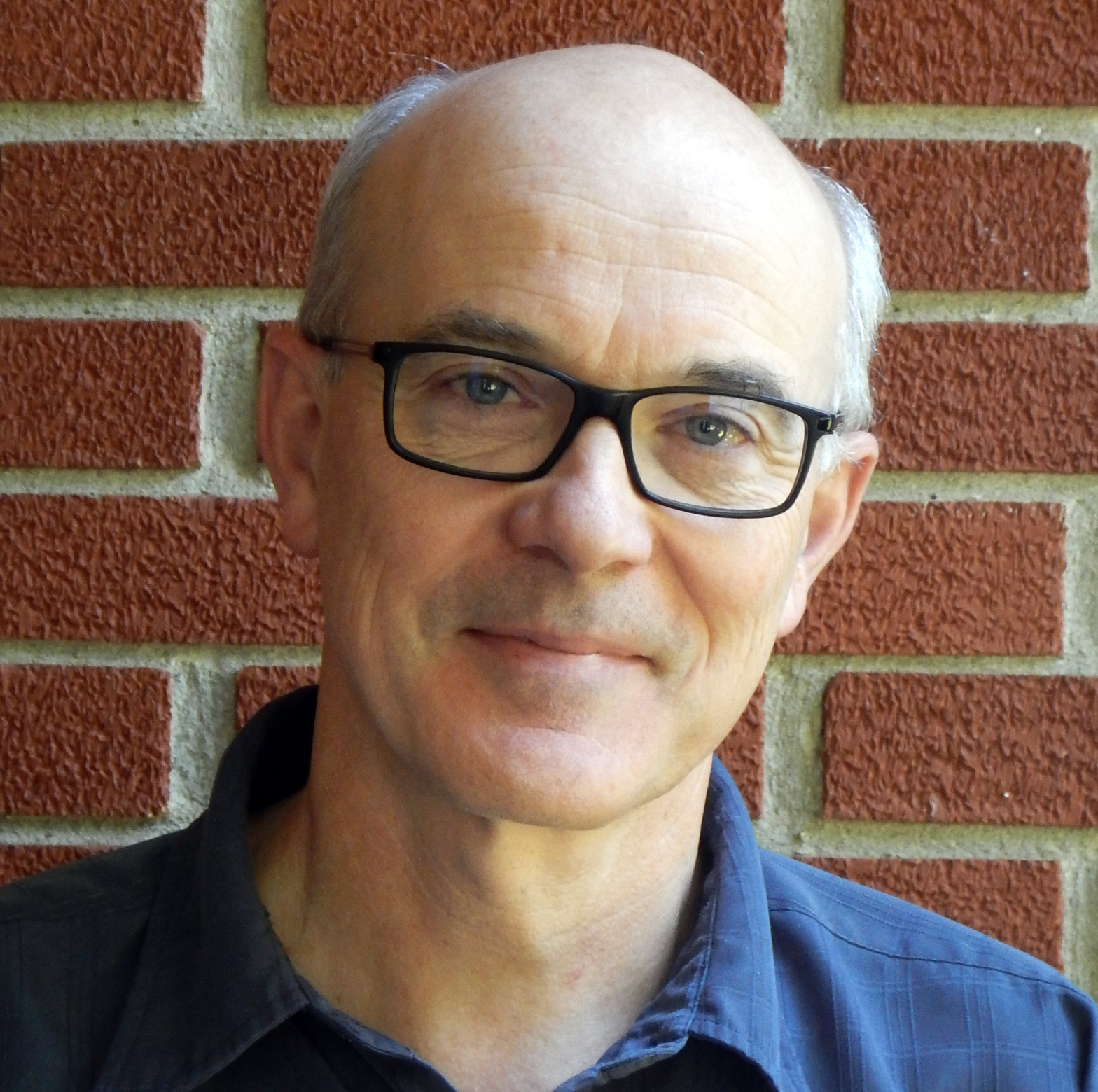 Close-up of a middle-aged man with glasses, short hair, wearing a dark blue shirt, standing in front of a red brick wall.