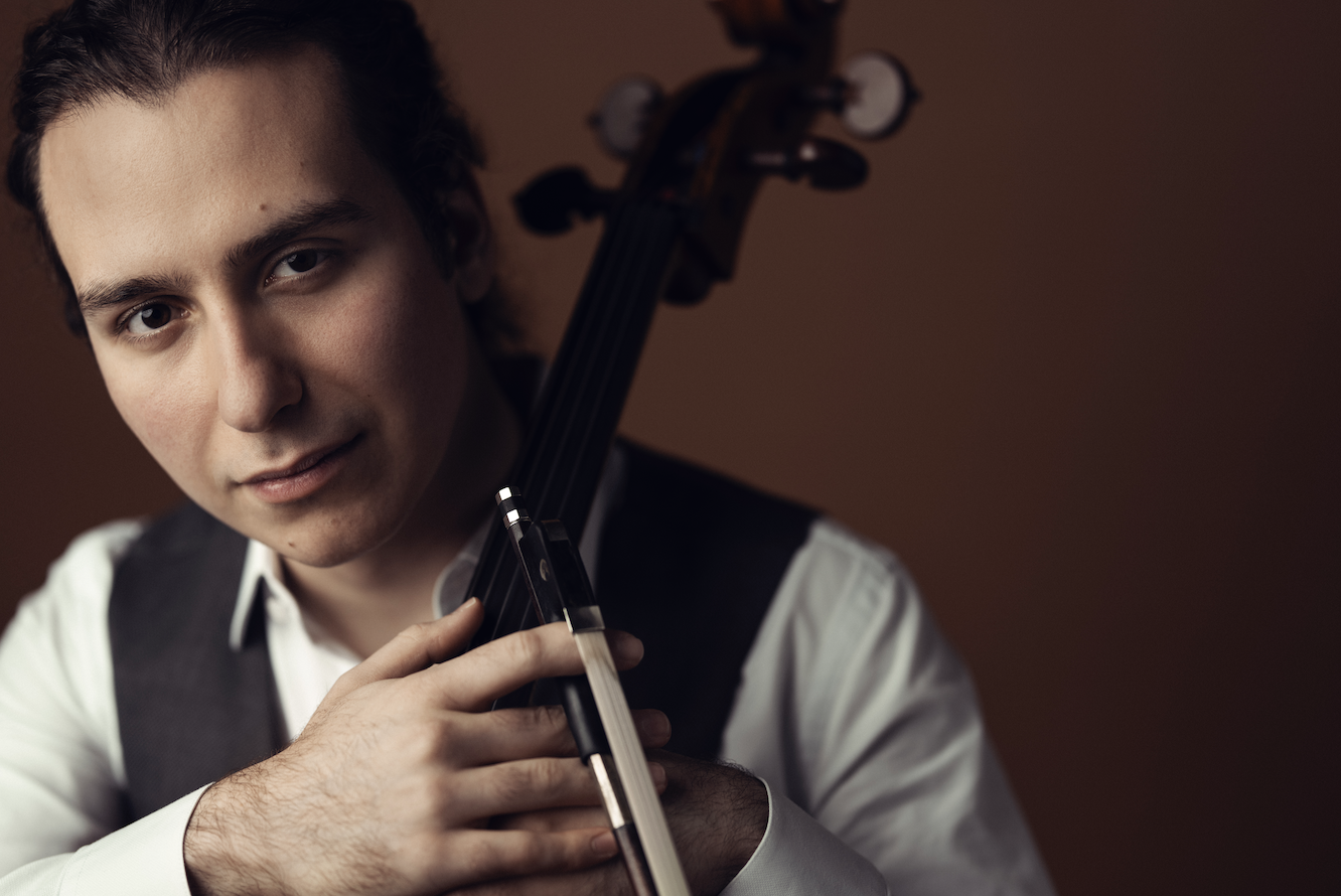 Young man with dark hair playing a violin, wearing a white shirt and vest, against a brown background.