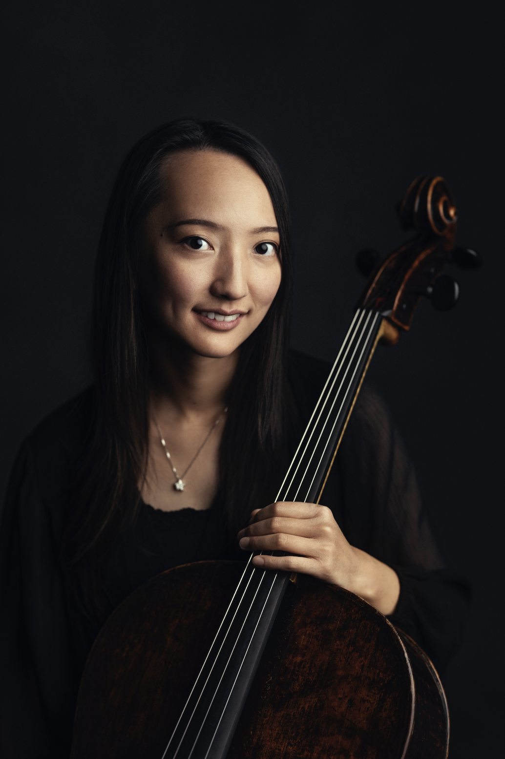 Portrait of an Asian woman with long dark hair, holding a cello, wearing a black top, and smiling against a dark background.