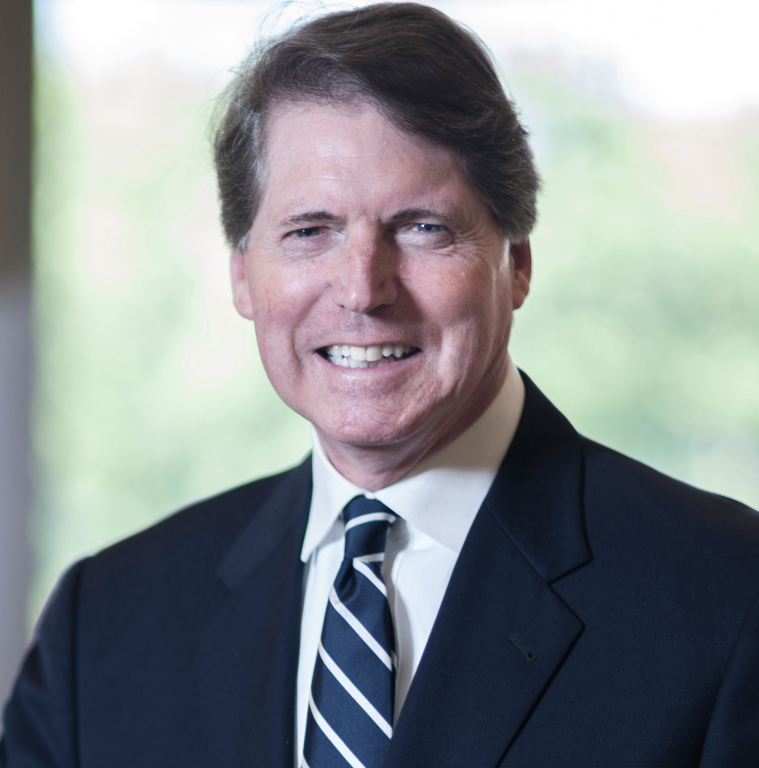 Portrait of a middle-aged man in a suit with a striped tie, smiling in front of a blurred natural background.
