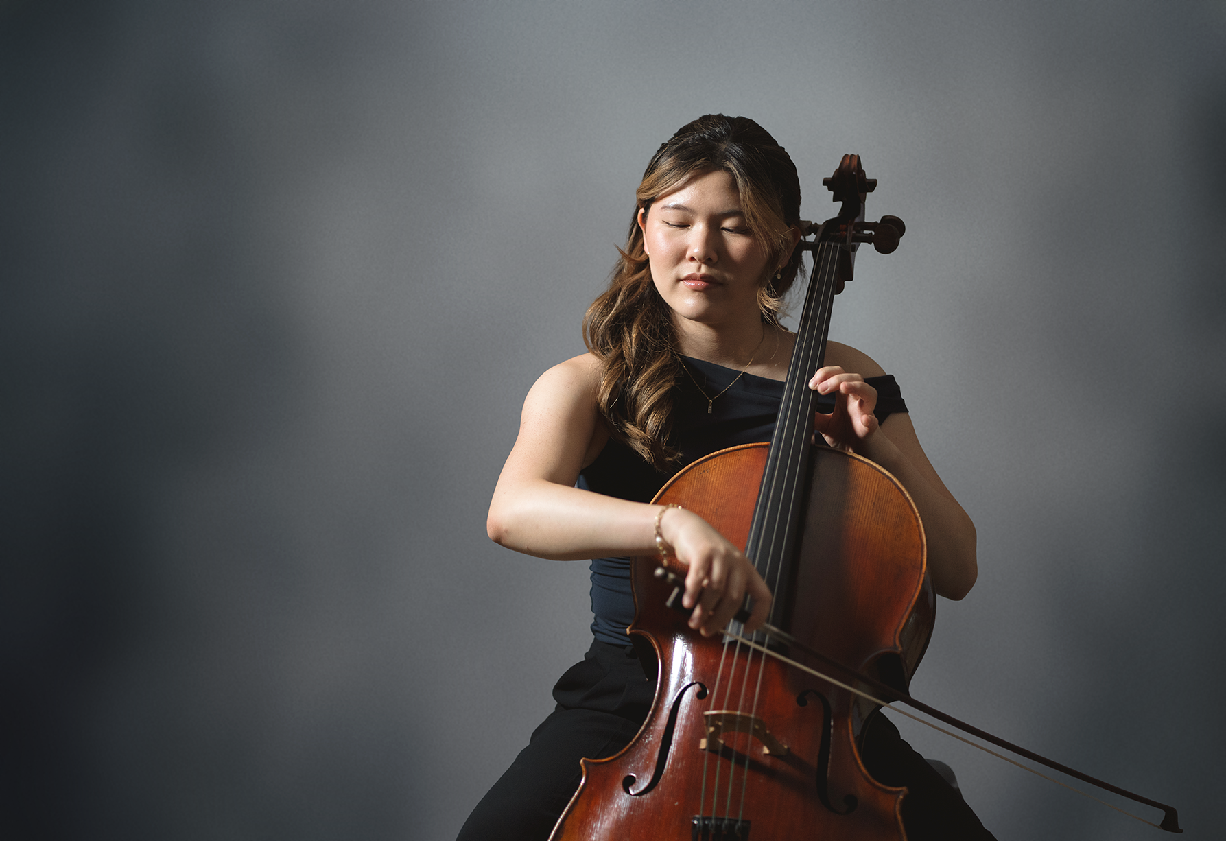 A woman with long wavy hair, wearing a black sleeveless top, plays the cello with eyes closed against a plain gray background.