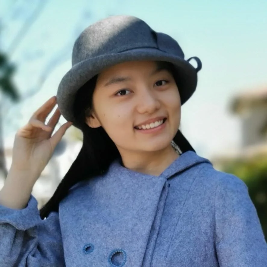 A young woman outdoors wearing a gray hat and blue hoodie, smiling and holding her hat with one hand.