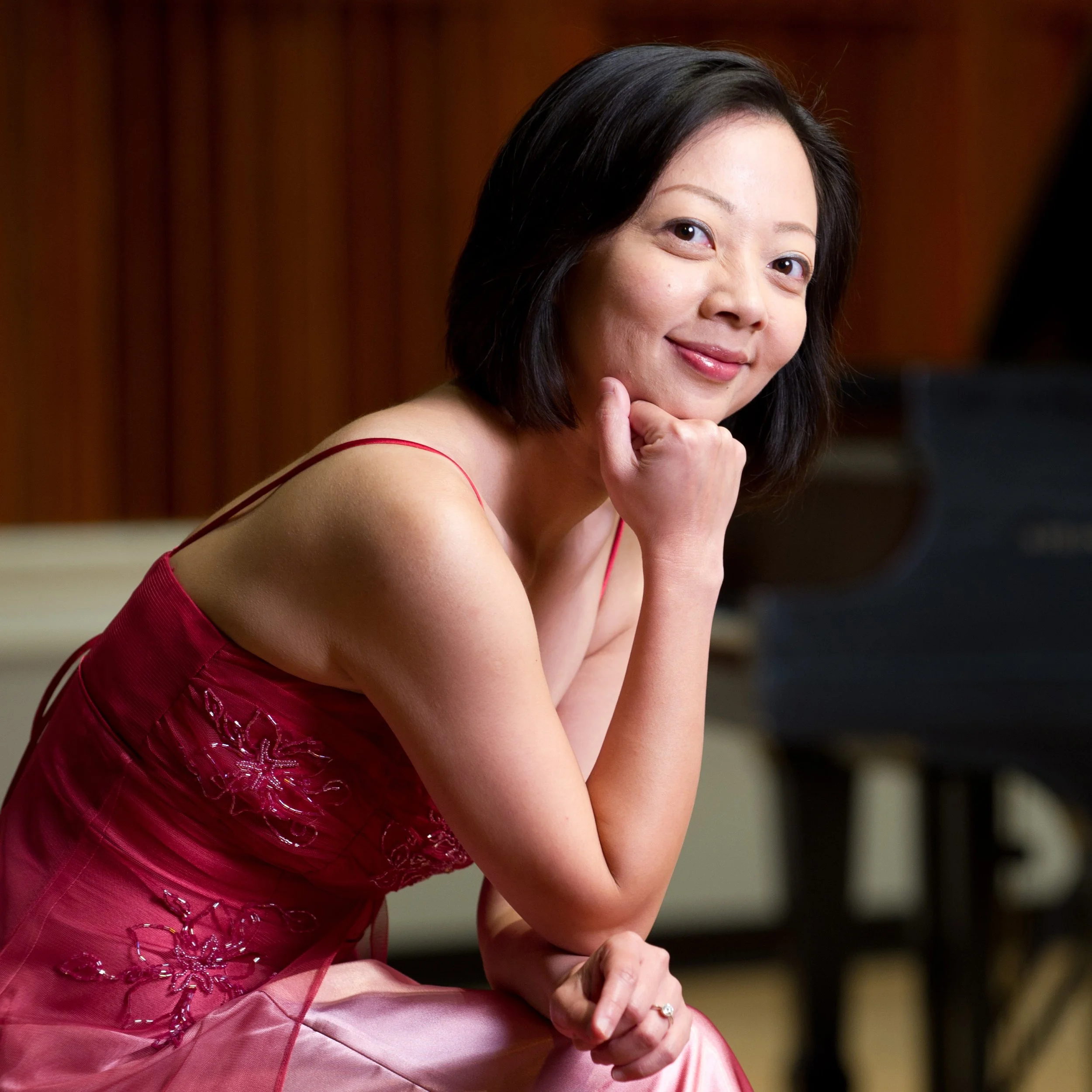 A woman in a pink dress sitting in front of a piano, smiling and resting her chin on her hand.