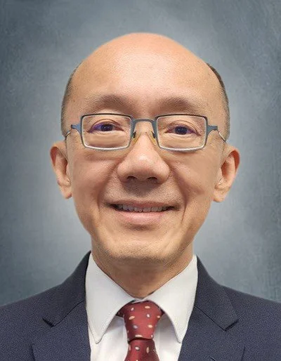 A man with glasses, short hair, wearing a dark suit, white shirt, and red polka dot tie, smiling against a plain gray background.