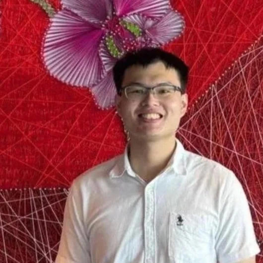 Young man wearing glasses and a white button-up shirt, smiling, standing in front of a red geometric-patterned background with a purple and green floral decoration.