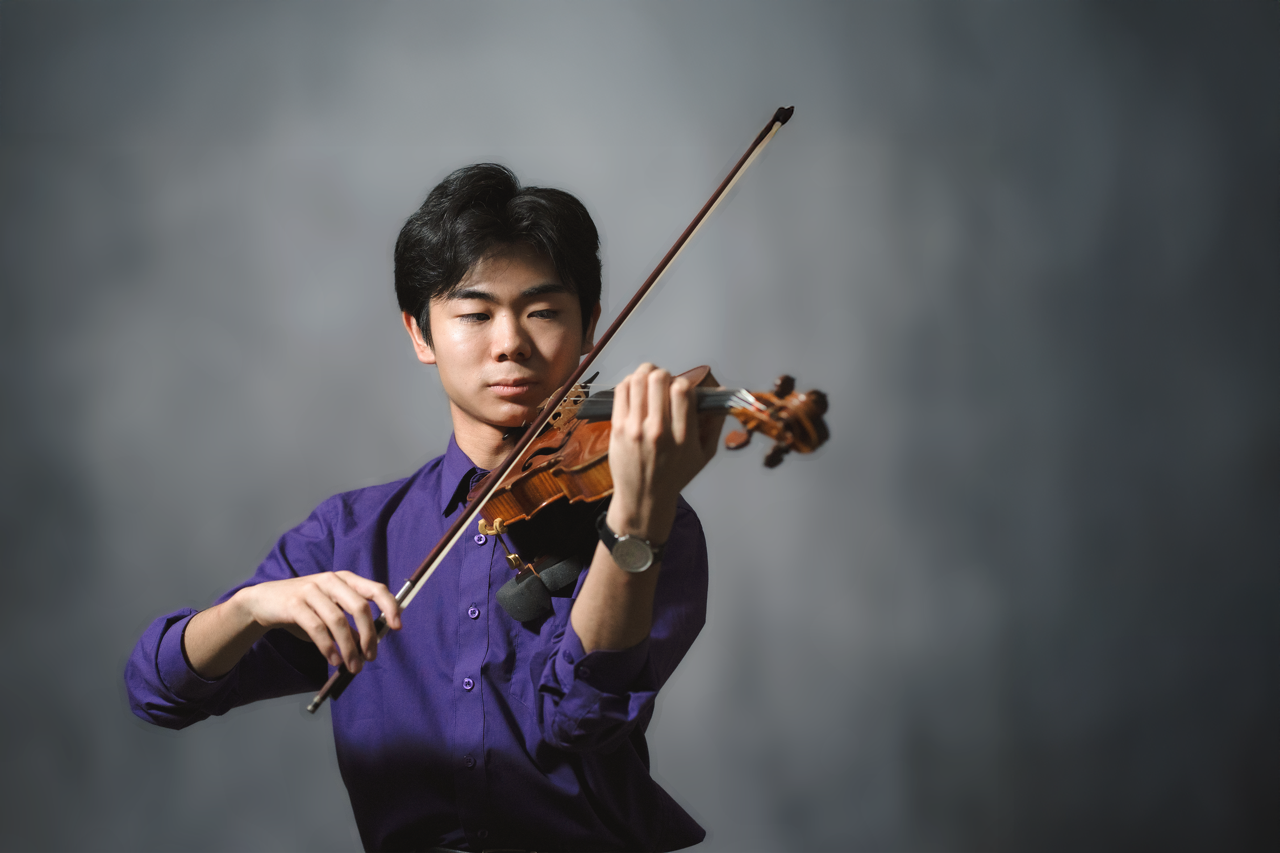 A young man with black hair, wearing a purple shirt, is playing a violin against a gray background.