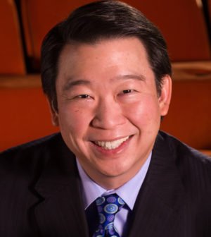 A man with short black hair, smiling, wearing a suit and a tie, in an indoor setting.
