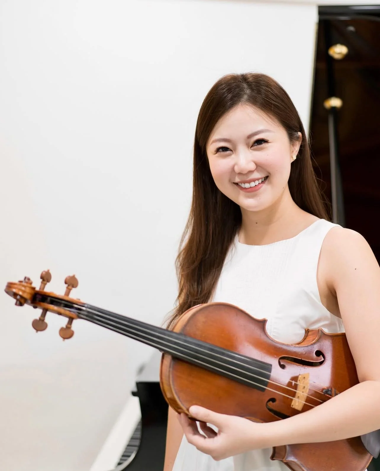 A smiling young woman holding a violin in a room with a piano.