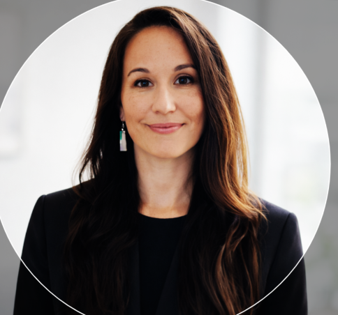 Professional woman with long brown hair, wearing a black blazer and earrings, smiling in a well-lit indoor setting.