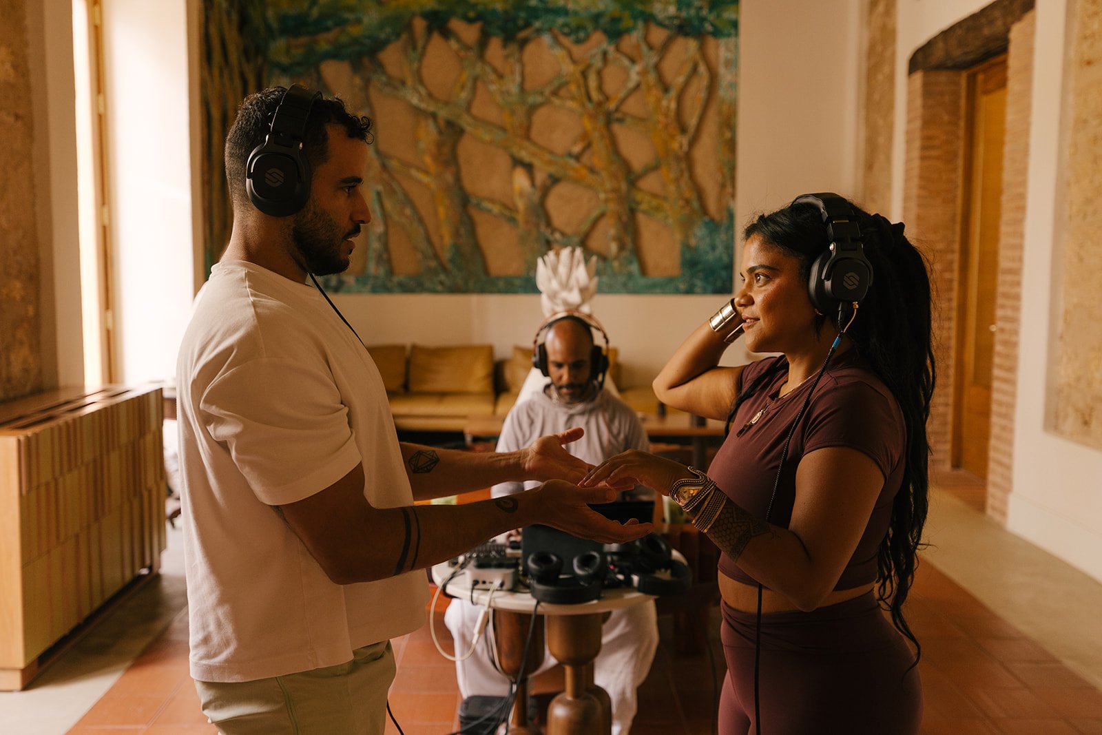 Two people in a recording studio, wearing headphones, facing each other and holding hands, with a man in the background at a mixing console.