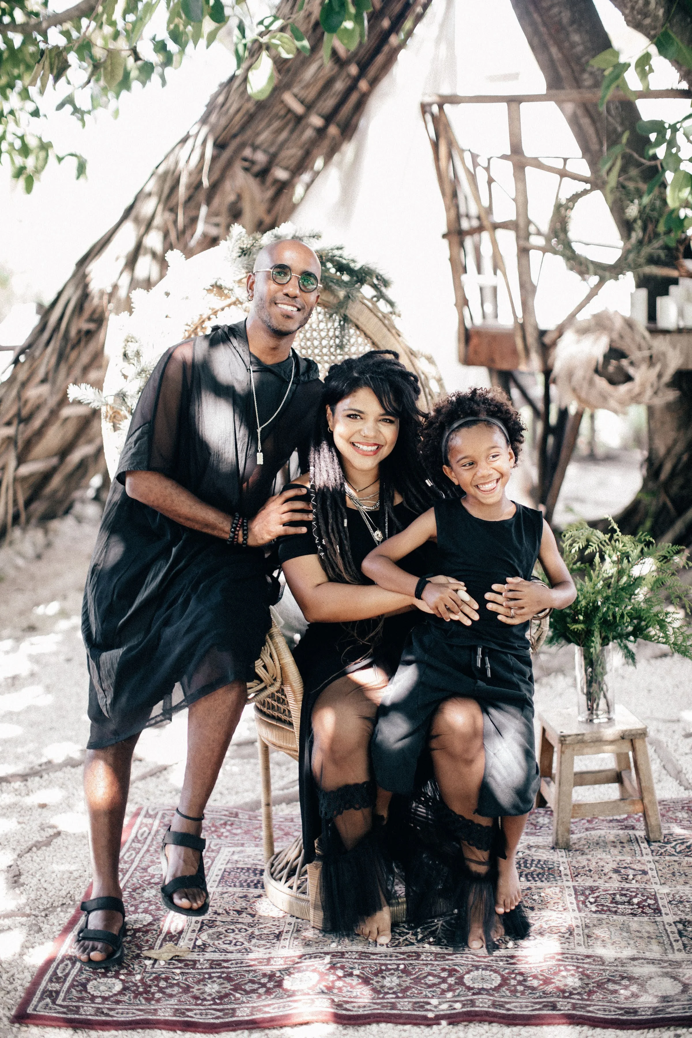 Three people, a man, woman, and girl, dressed in black, smiling and posing for a photo outdoors, with a rustic wooden backdrop, a small table with a plant, and a patterned rug underneath them.