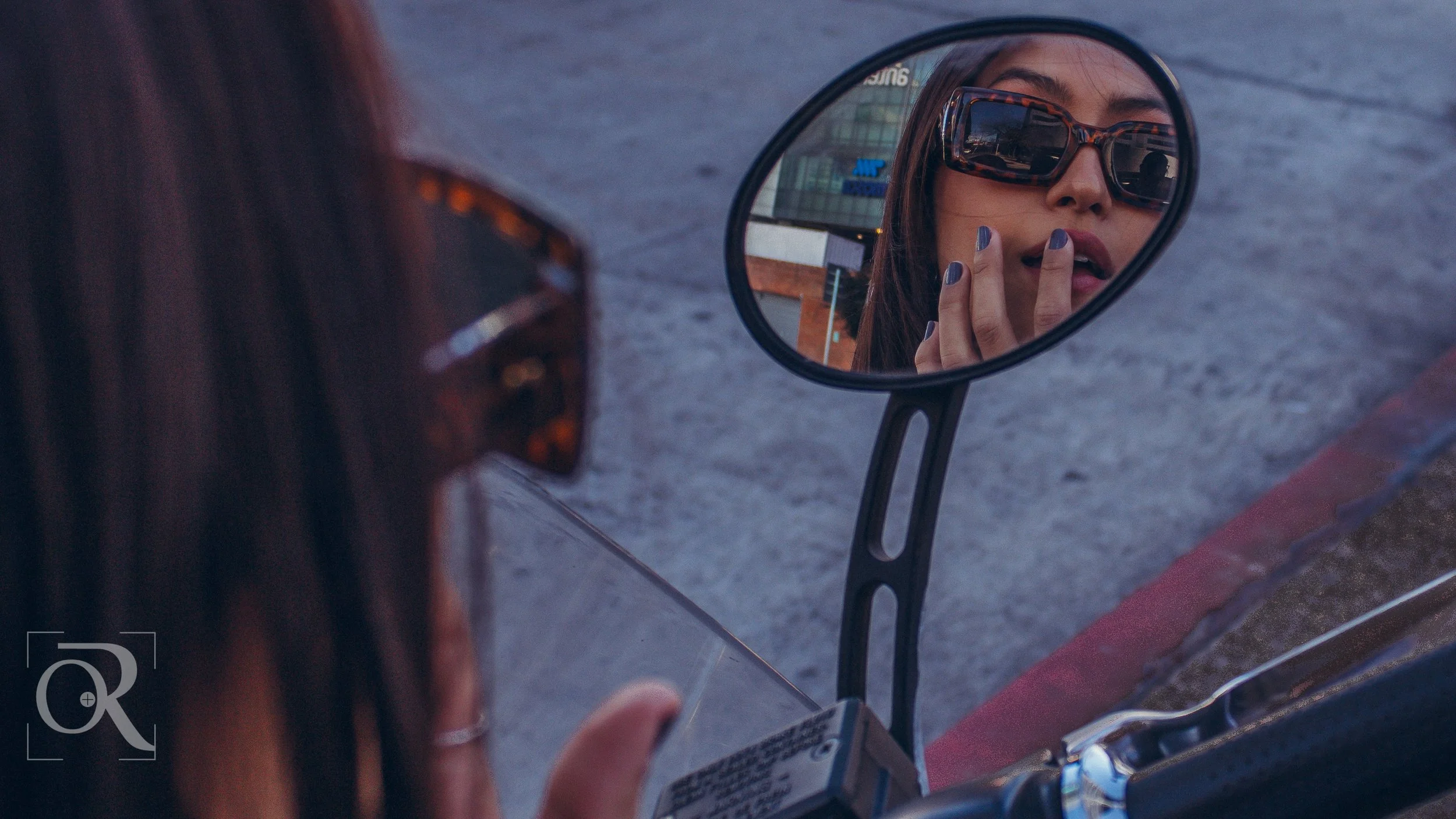 Mujer con gafas de sol mirándose en el espejo de una motocicleta, tocándose los labios con la mano.