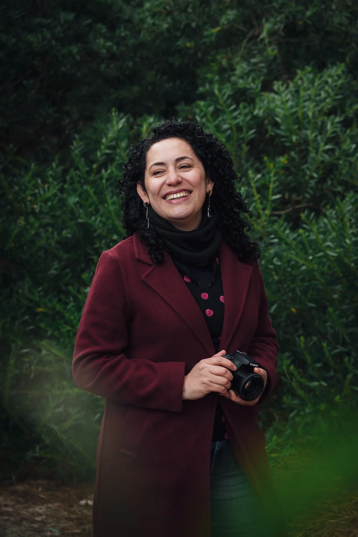Mujer sonriendo con cámara en un entorno natural con vegetación verde.