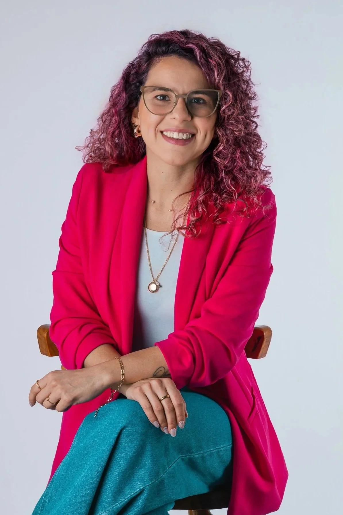 Mujer con cabello rizado y teñido de rosa, gafas, blazer rosa y pantalón azul, sonriente y posando en estudio.