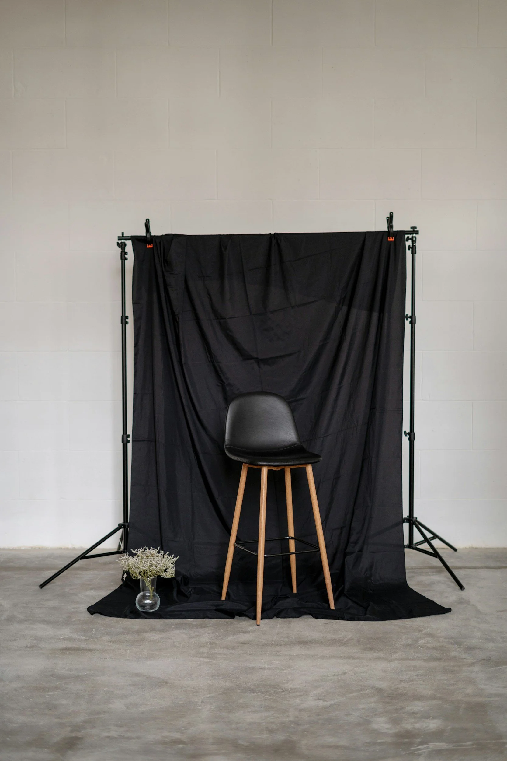 Una silla negra con patas de madera frente a un fondo negro sobre un soporte, acompañado de un florero con flores blancas en la esquina inferior izquierda.