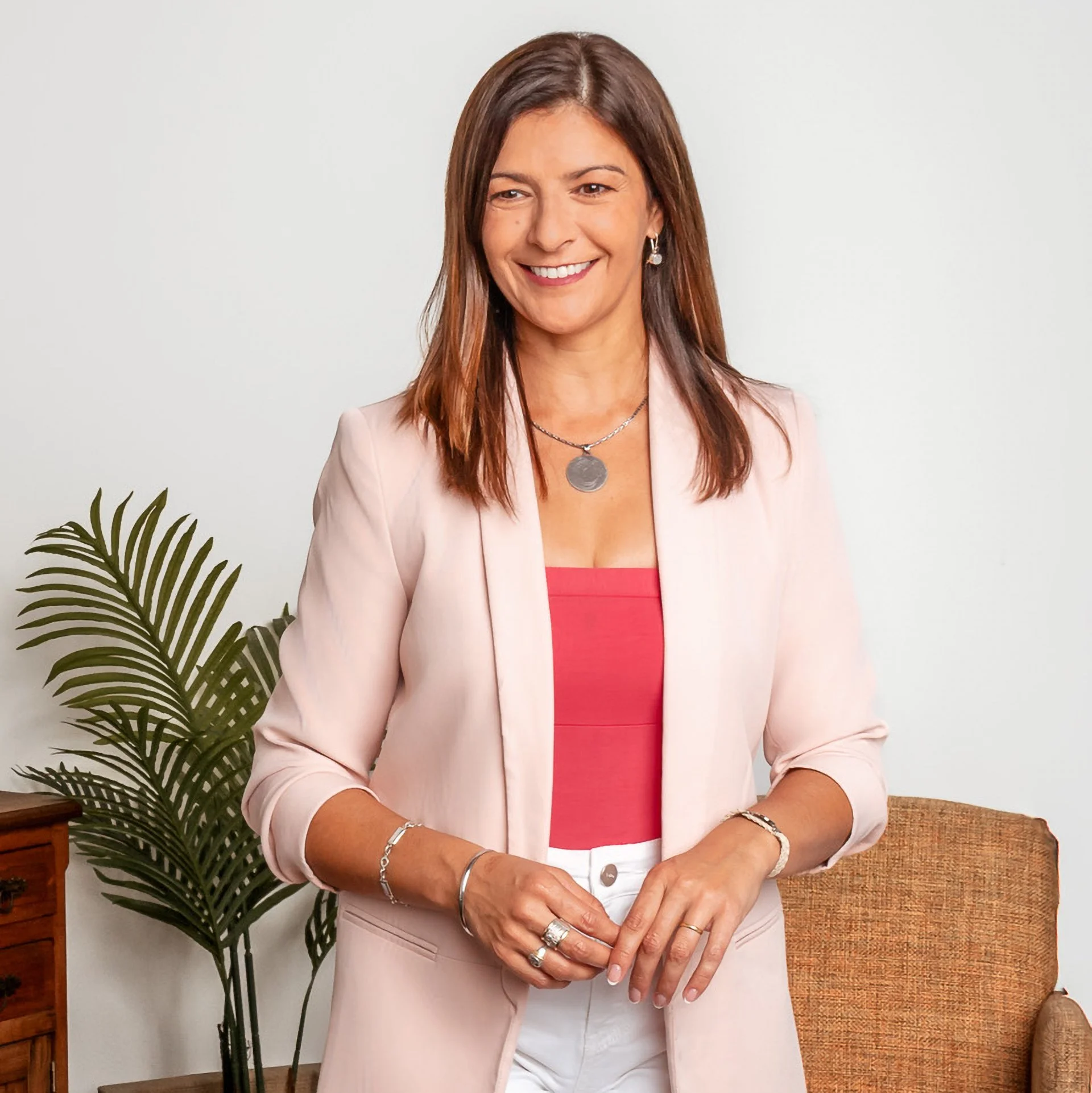 Mujer sonriendo con blusa rosa y blazer en un ambiente interior con planta y sillón.